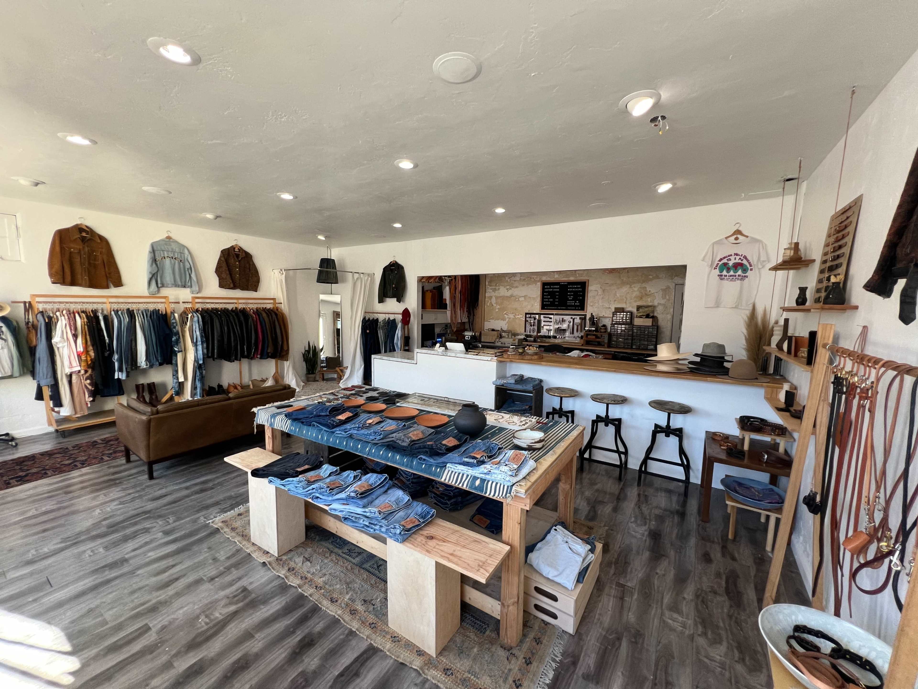 An interior of a clothing store featuring racks of apparel, a central display table with denim items, and a minimalist wooden counter area.
