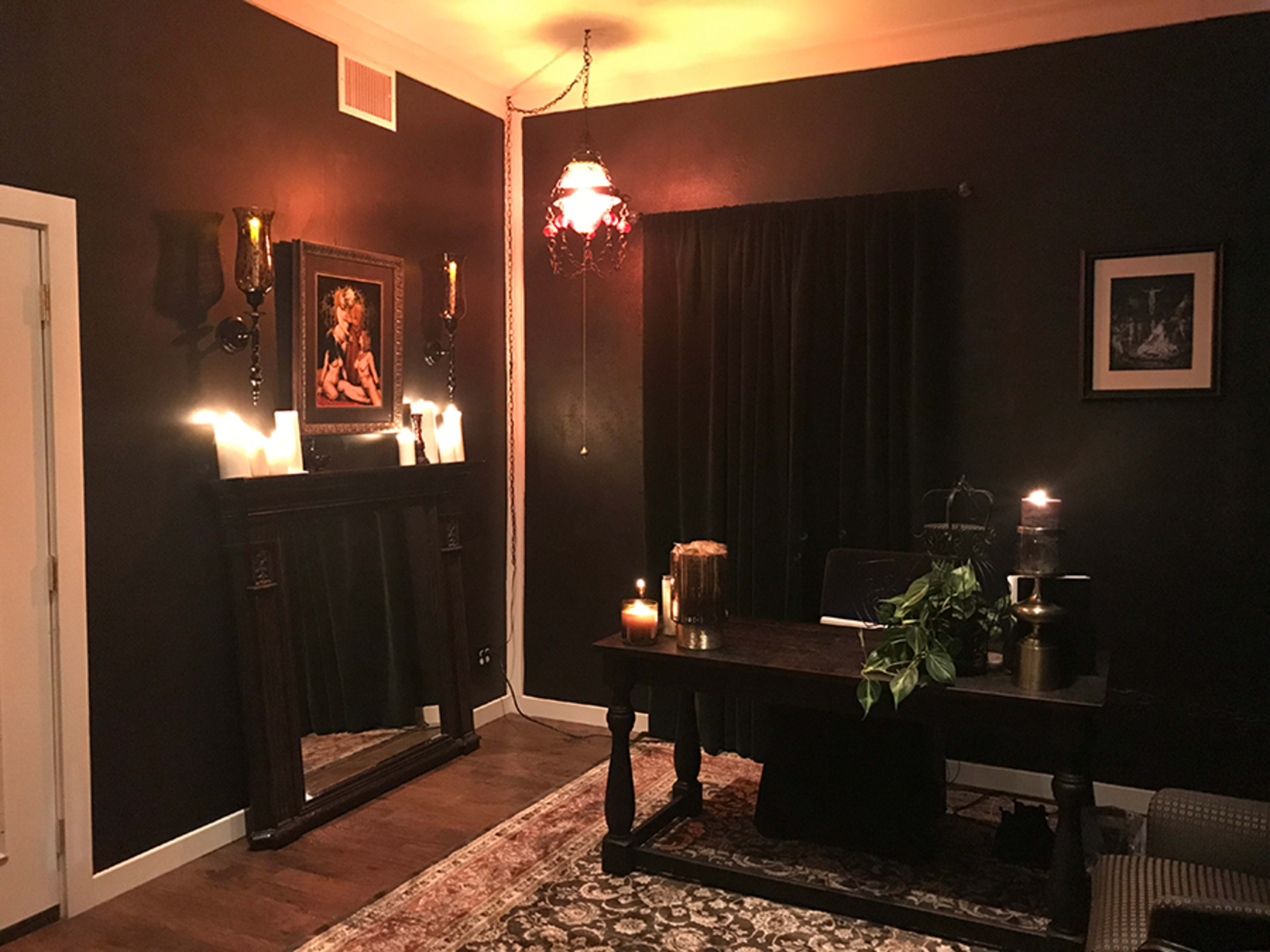 A dimly lit room featuring a dark-colored wall, a wooden desk with candles, a mirror, and a framed picture, all arranged around an area rug.