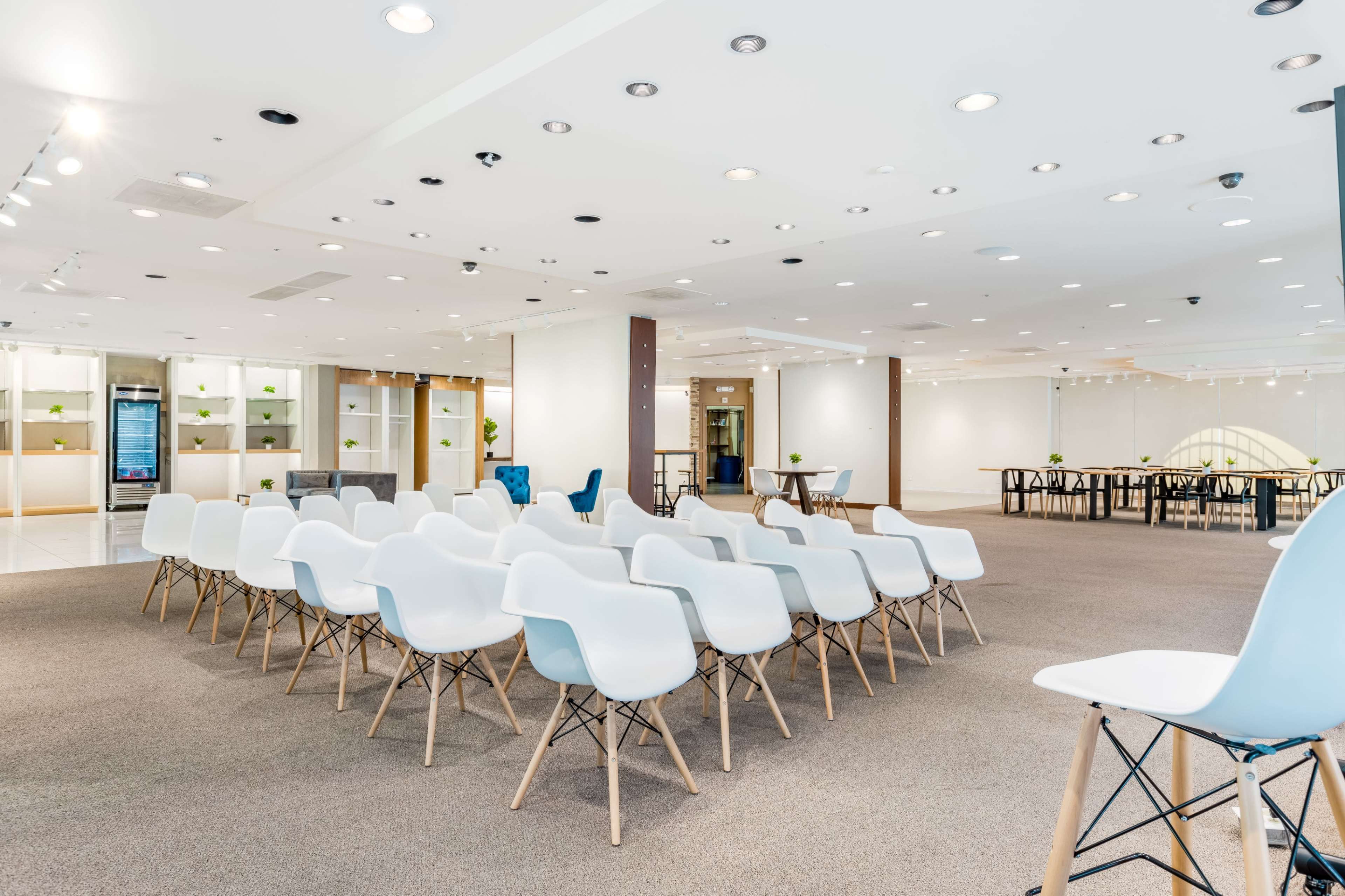A spacious, modern conference room with rows of white chairs set up for an event, surrounded by minimalistic furniture and soft lighting.