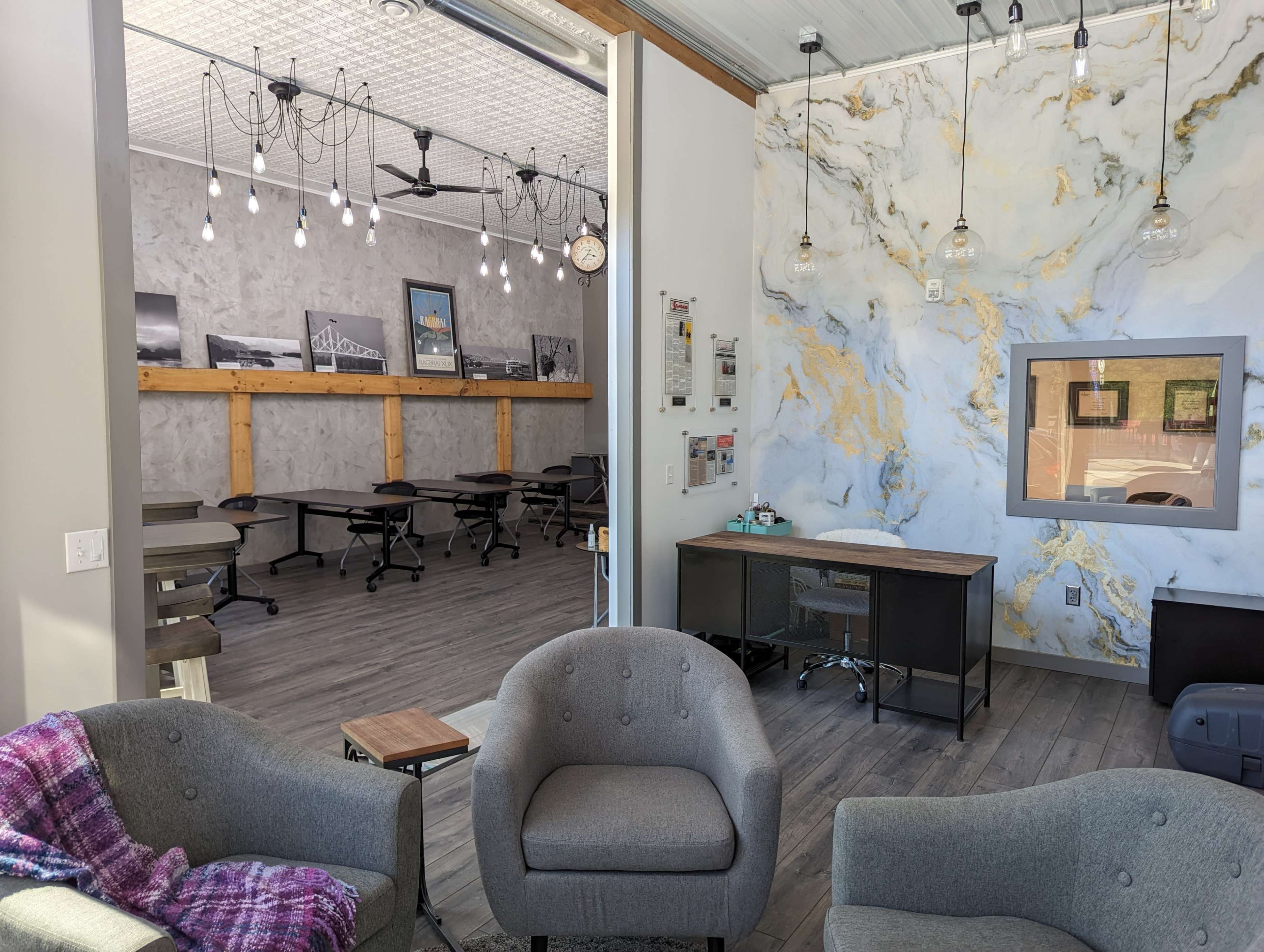 A modern workspace featuring a seating area with gray chairs and a wooden table, leading to an office space with desks, artwork on the walls, and overhead lights.