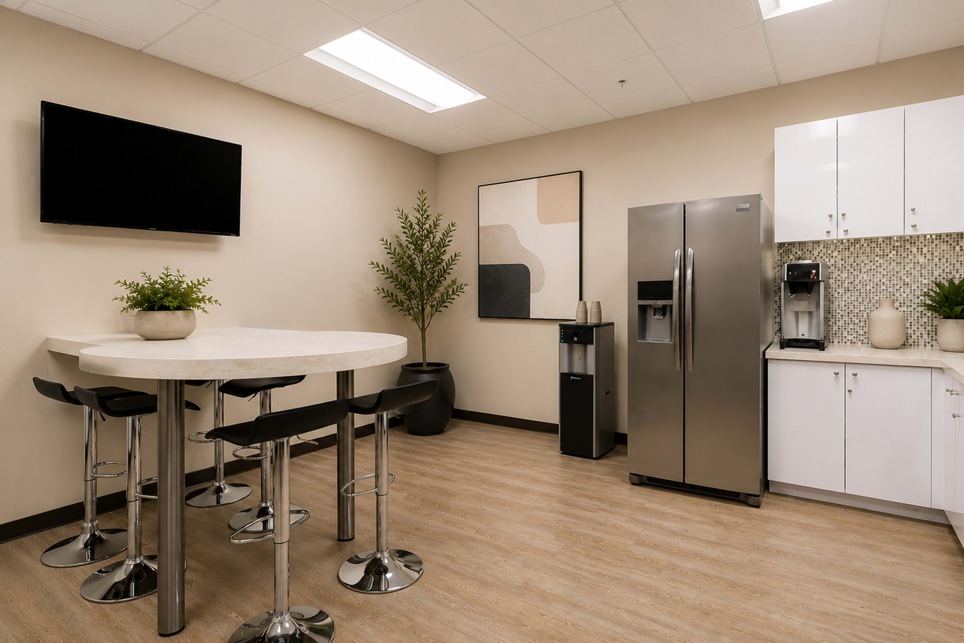 The image shows a modern break room featuring a round table with high chairs, a refrigerator, a coffee machine, and decorative plants.