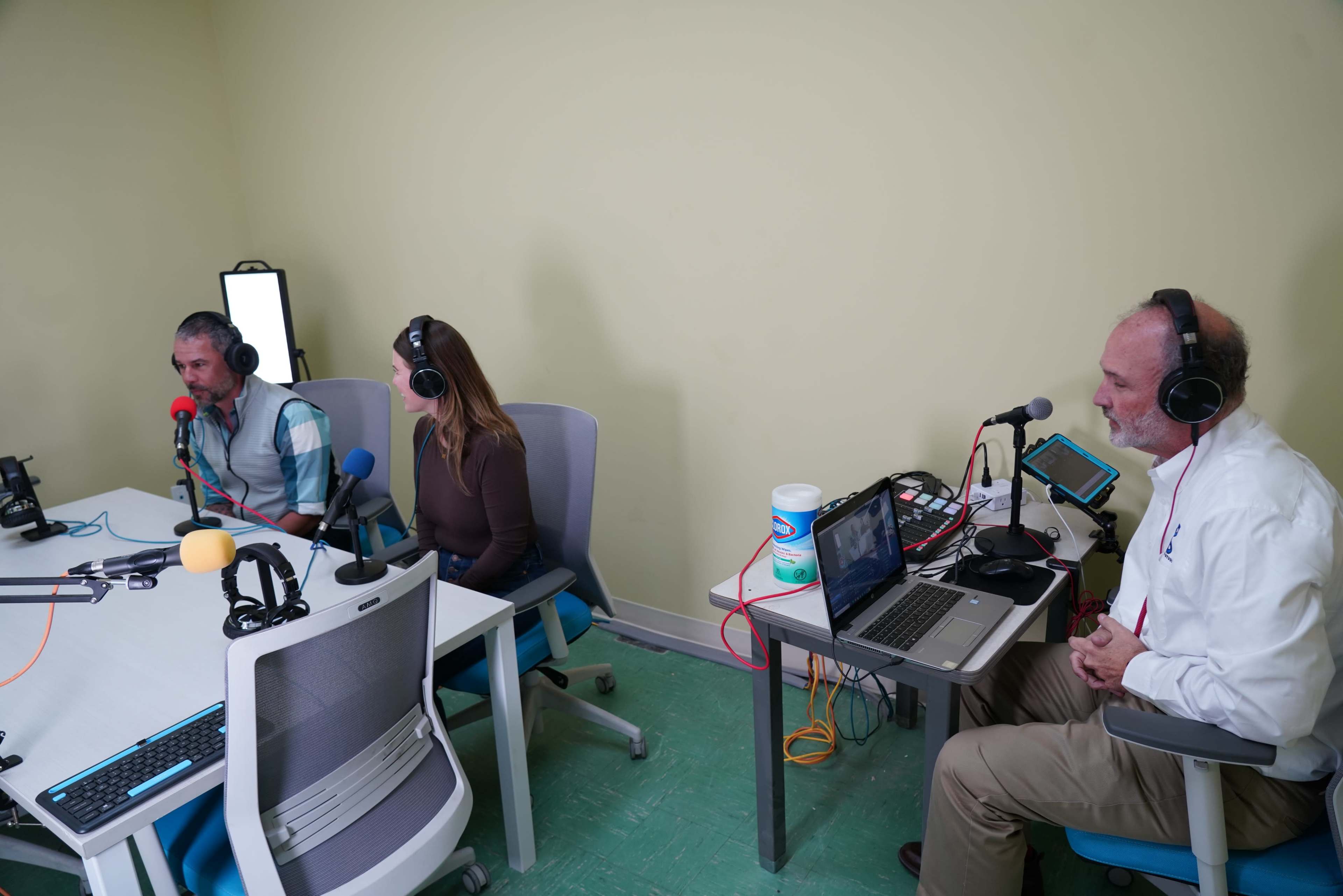 Three individuals are seated at a table with microphones and equipment in a small recording studio.