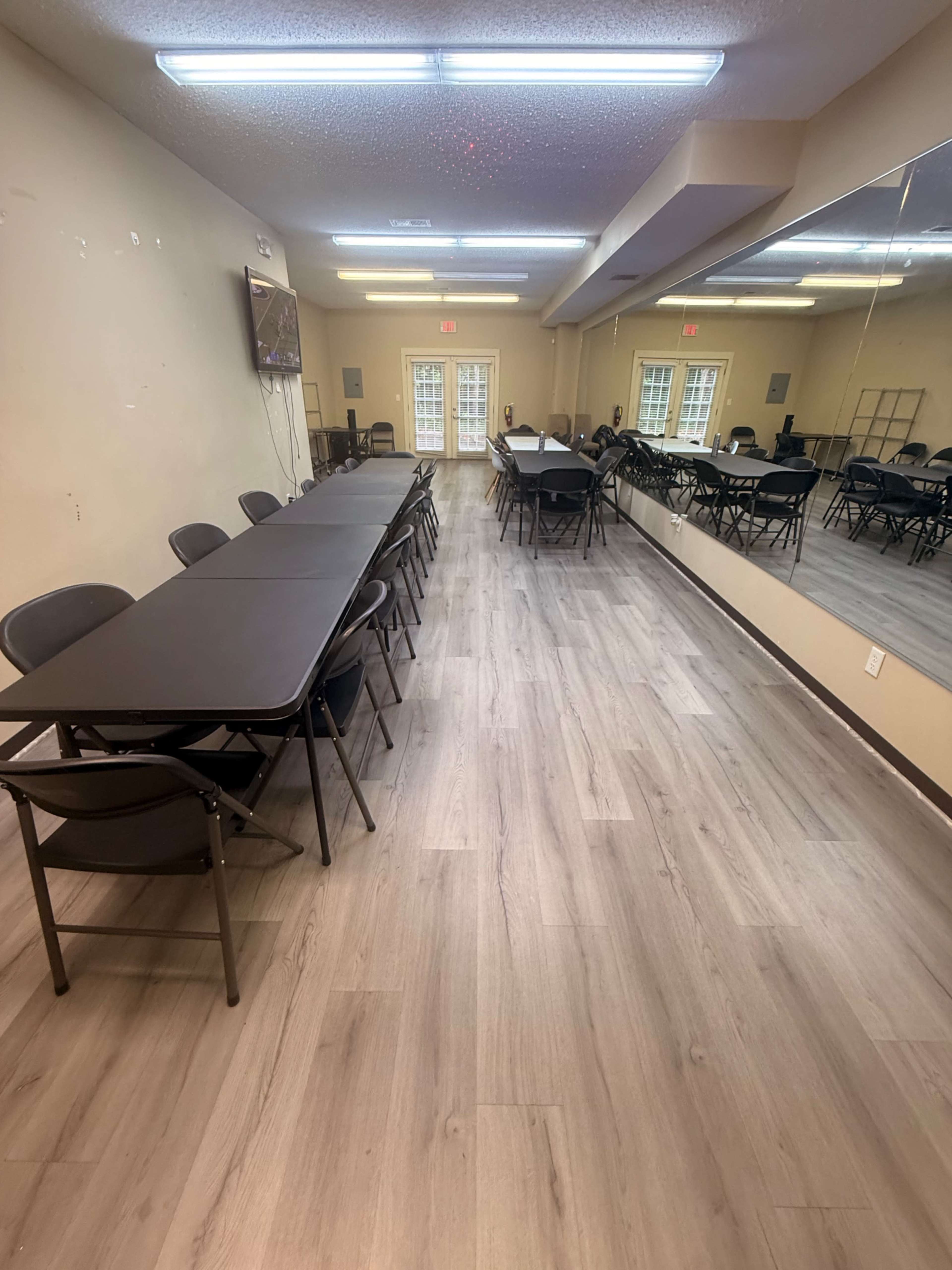 The image shows a spacious, well-lit meeting room with several rows of folding tables and chairs arranged systematically along with a mirror on one wall.