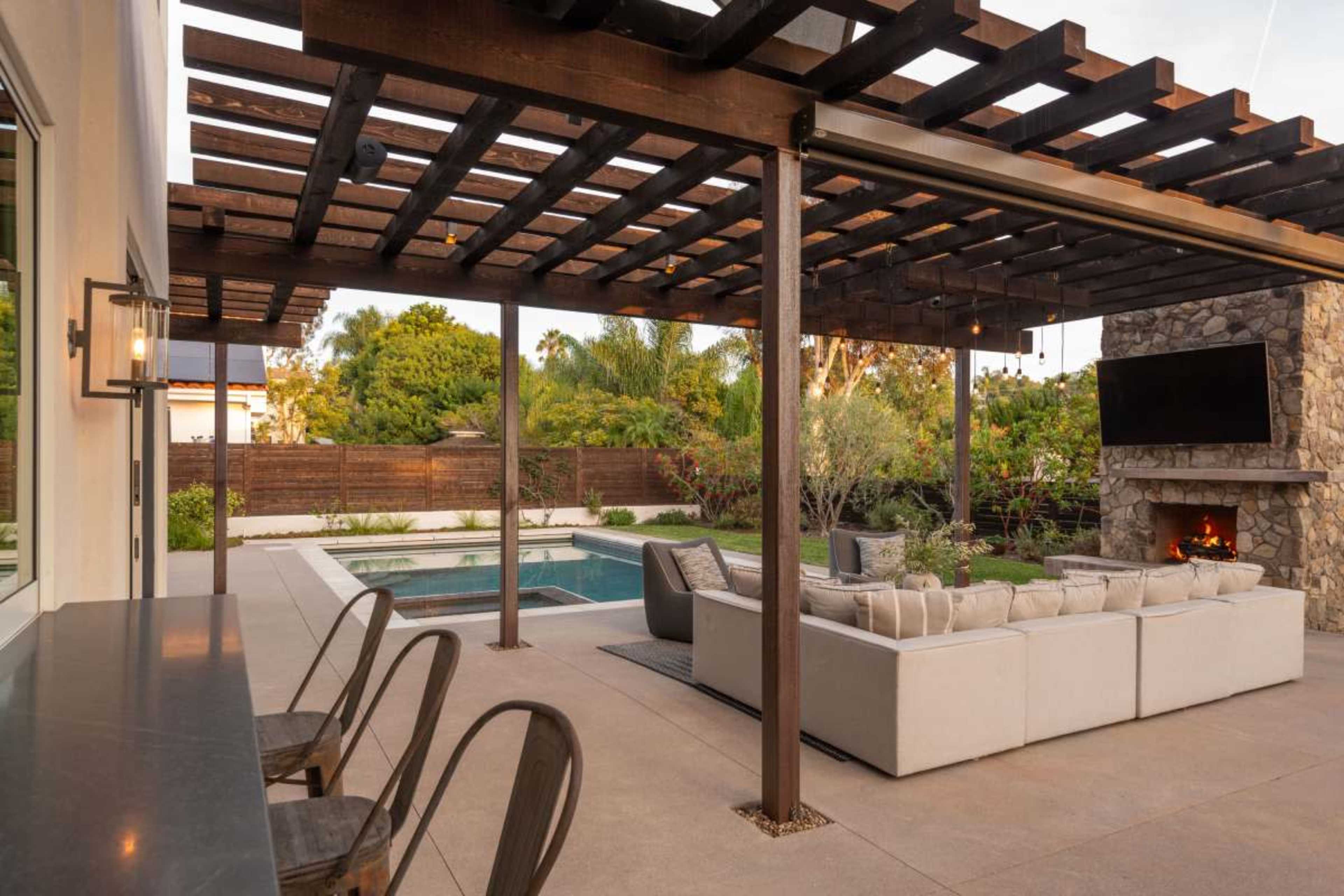 The image shows a spacious outdoor patio area with a stone fireplace, a swimming pool, and a sofa set beneath a wooden pergola.