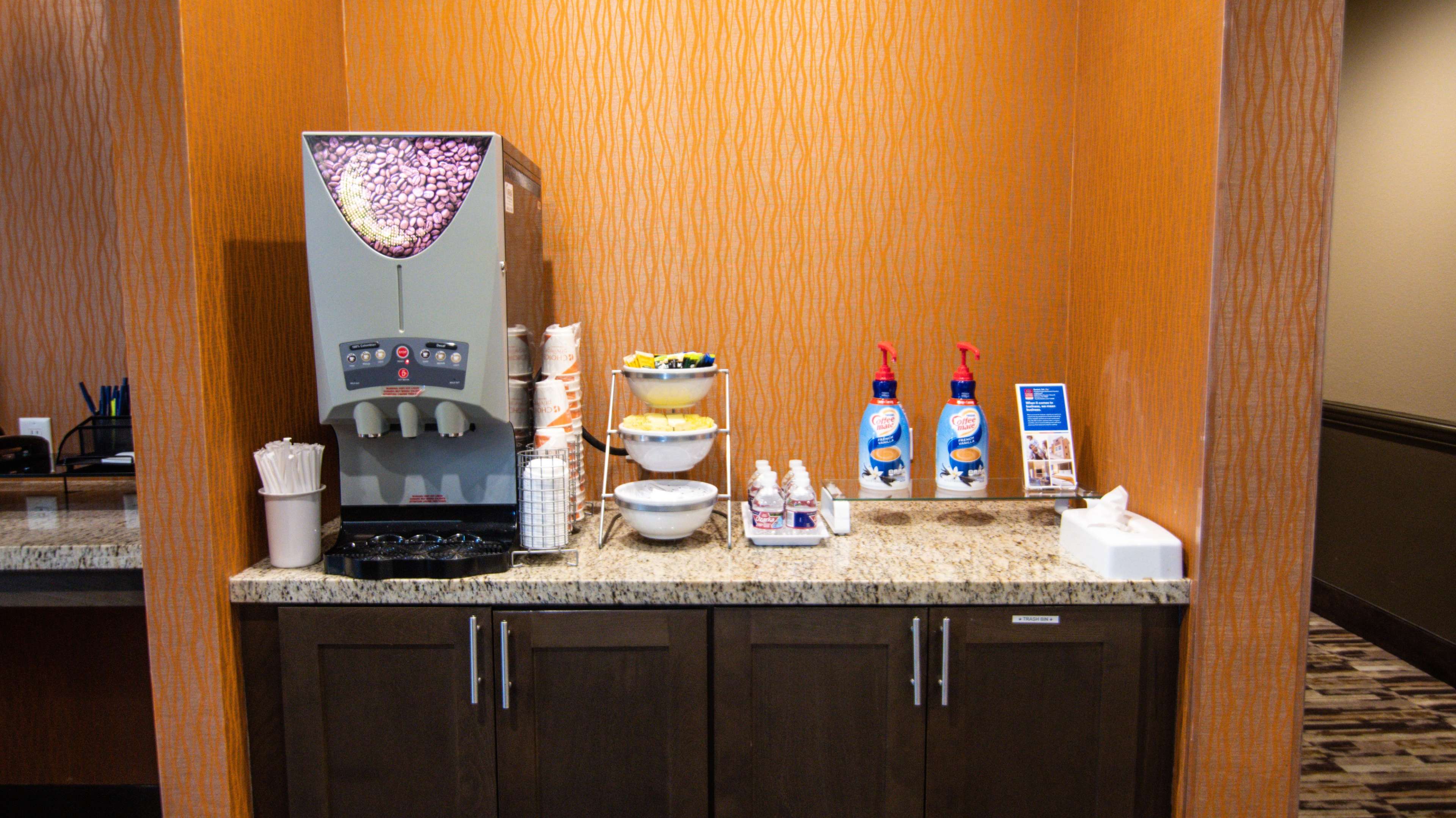 A beverage station featuring a soft serve ice cream machine, a bowl of toppings, dispensers of whipped cream, and assorted utensils on a countertop against an orange textured wall.