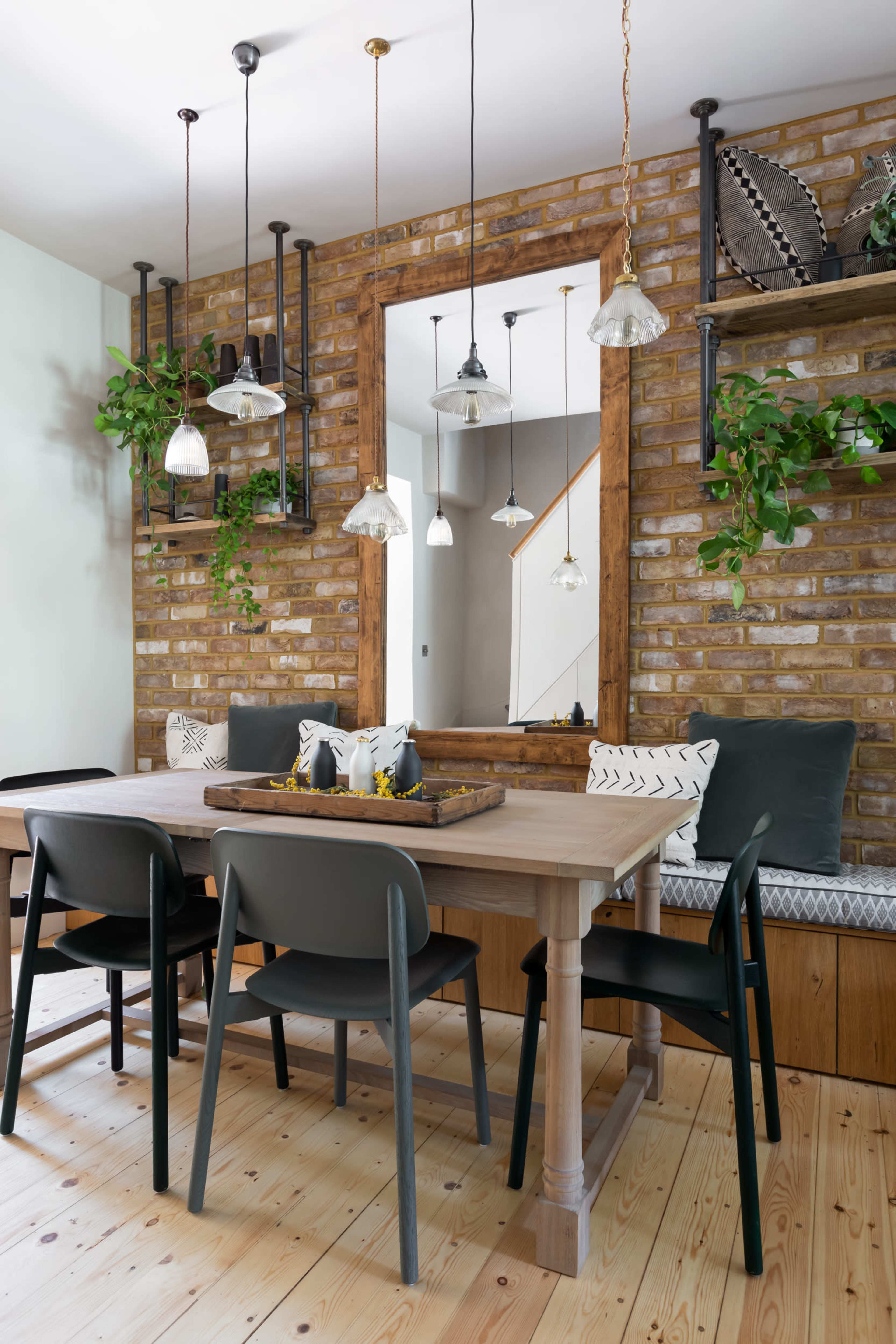 A wooden dining table surrounded by black chairs is positioned in a room with a brick wall and hanging pendant lights, featuring plants on shelves and a mirrored frame.