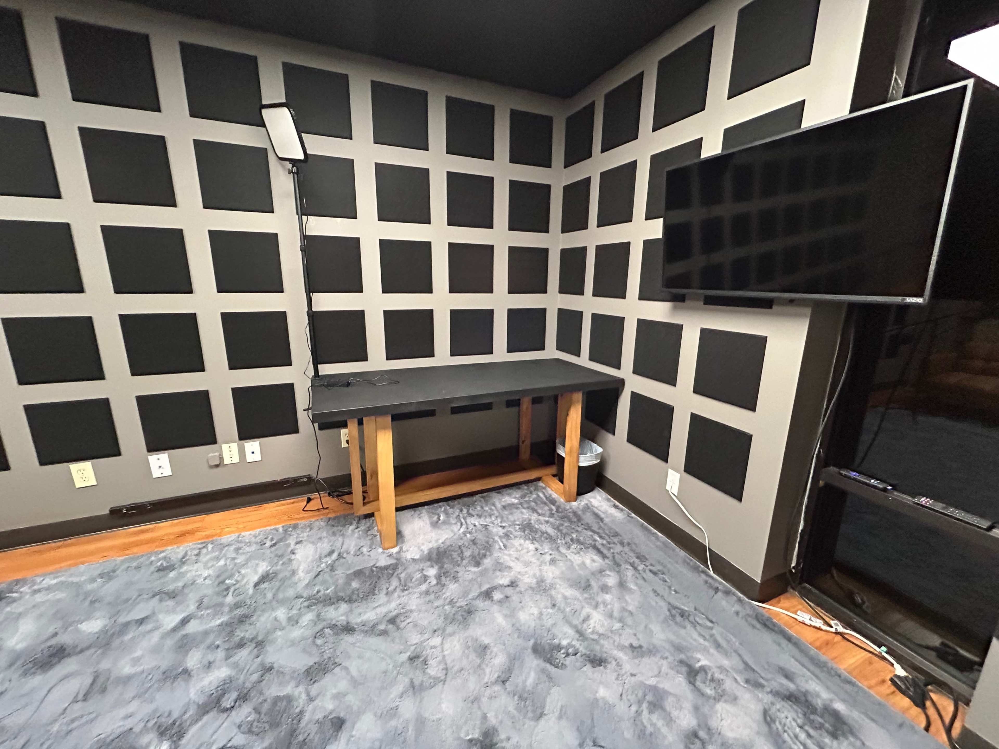 A room with textured black walls, a wooden desk in the center, and a flat-screen television mounted on the wall.