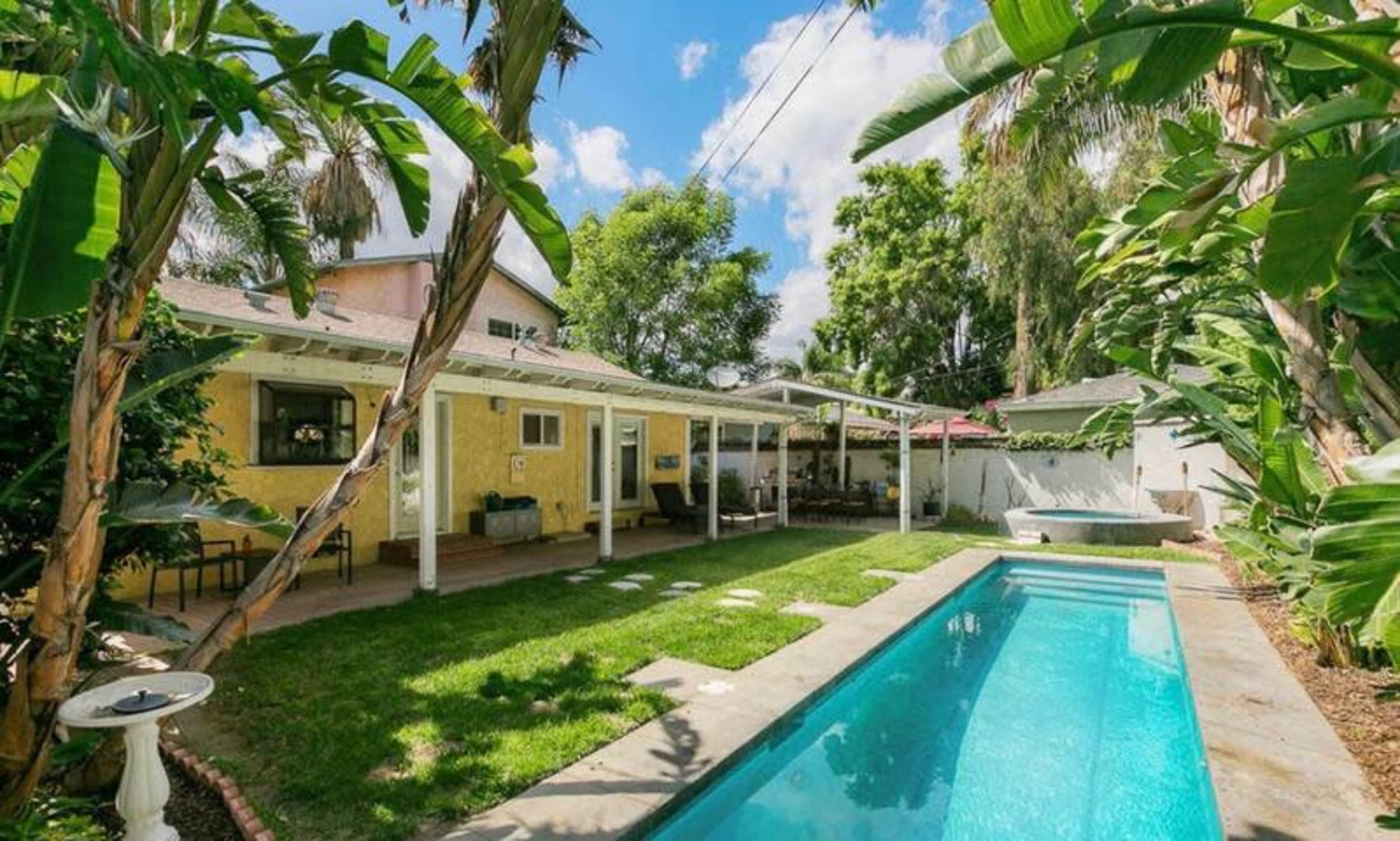 A backyard with a swimming pool, green lawn, and a covered patio area beside a house surrounded by trees and tropical plants.