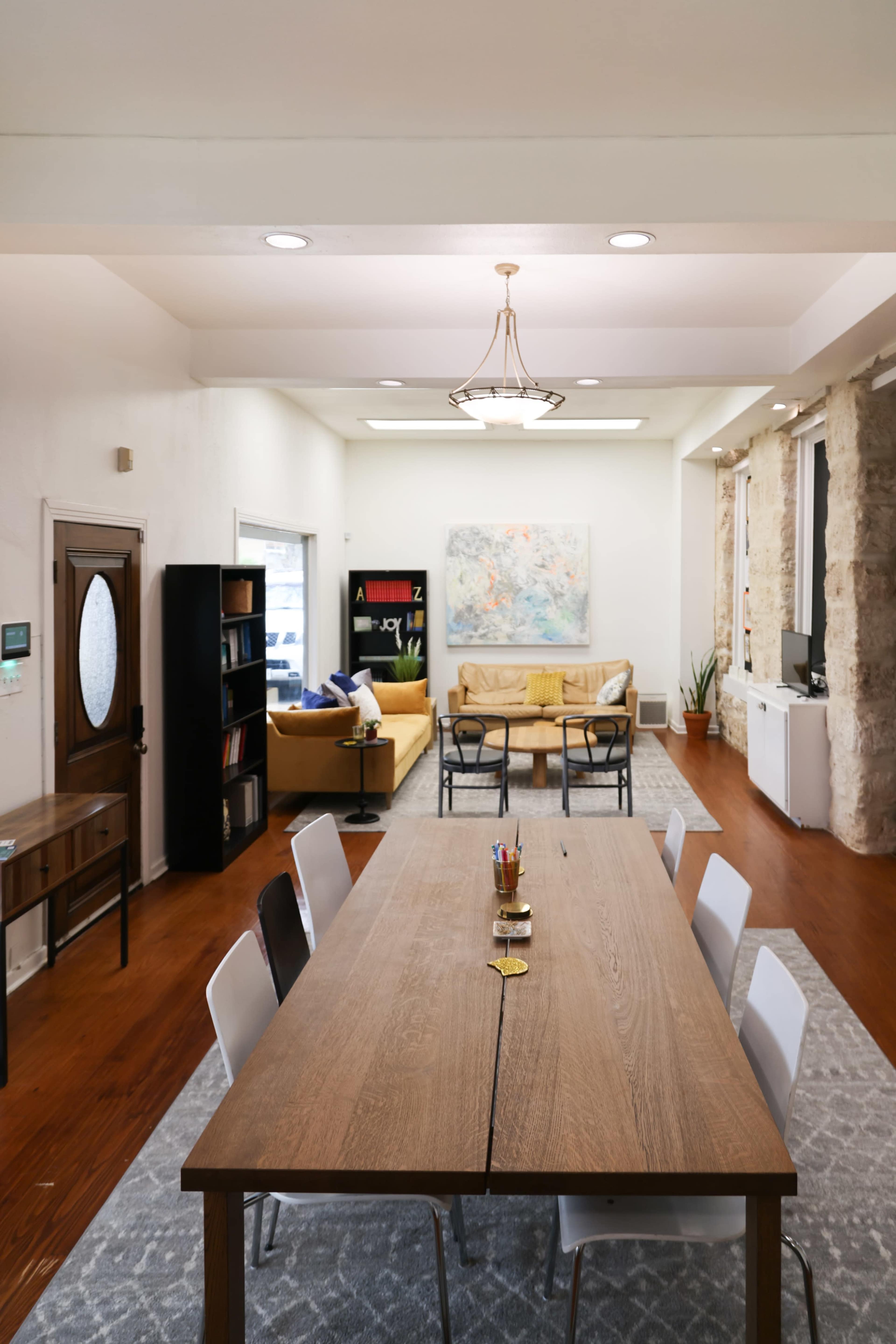 A modern open-concept workspace features a large wooden dining table in the foreground, with a seating area and artwork visible in the background.