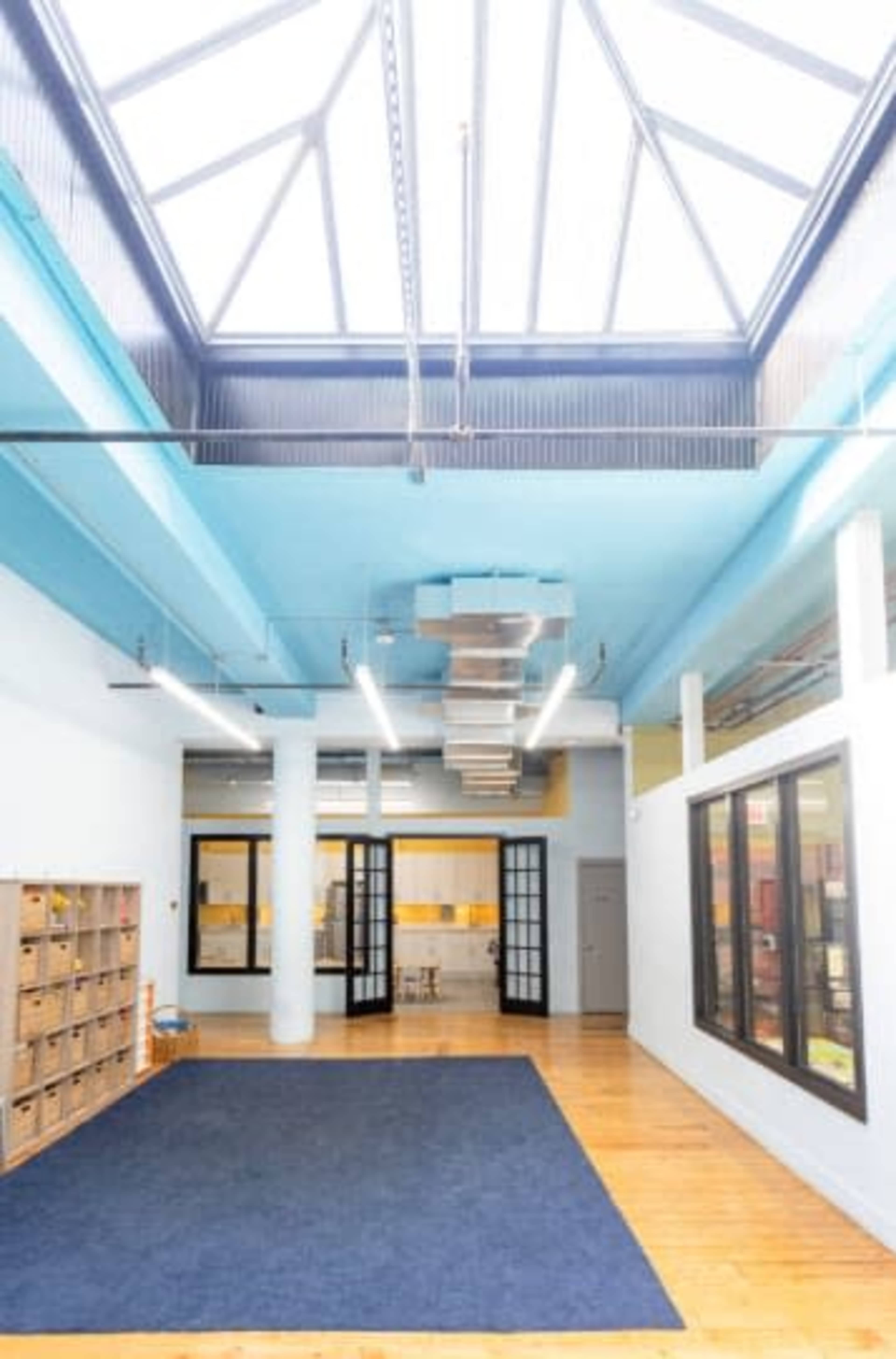 The image shows a bright, open space with a blue ceiling, large windows, a wooden floor, and a wall of cubby storage.