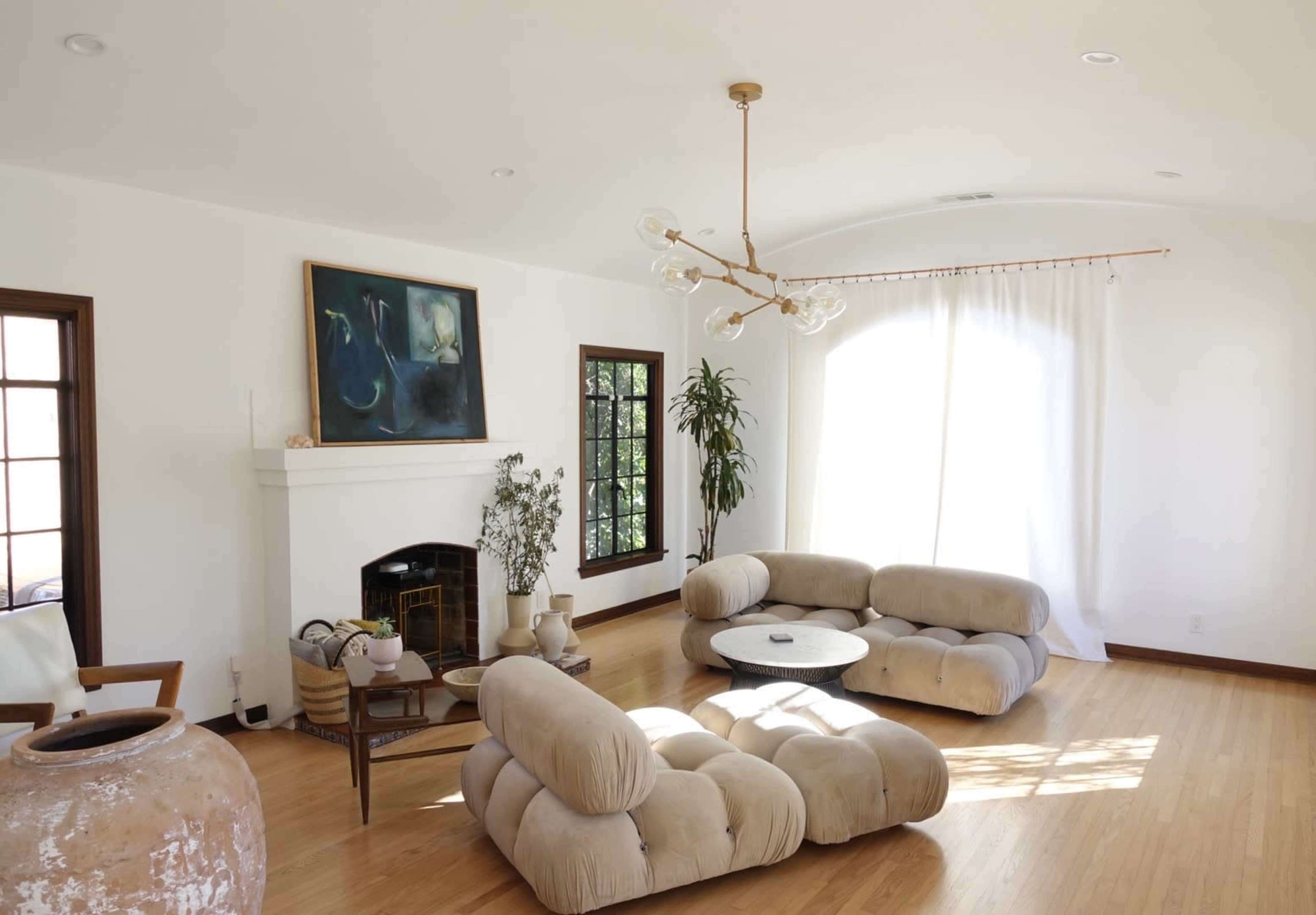 A spacious, well-lit living room features a unique, rounded beige sofa set, a circular coffee table, a fireplace, and large windows with sheer curtains.