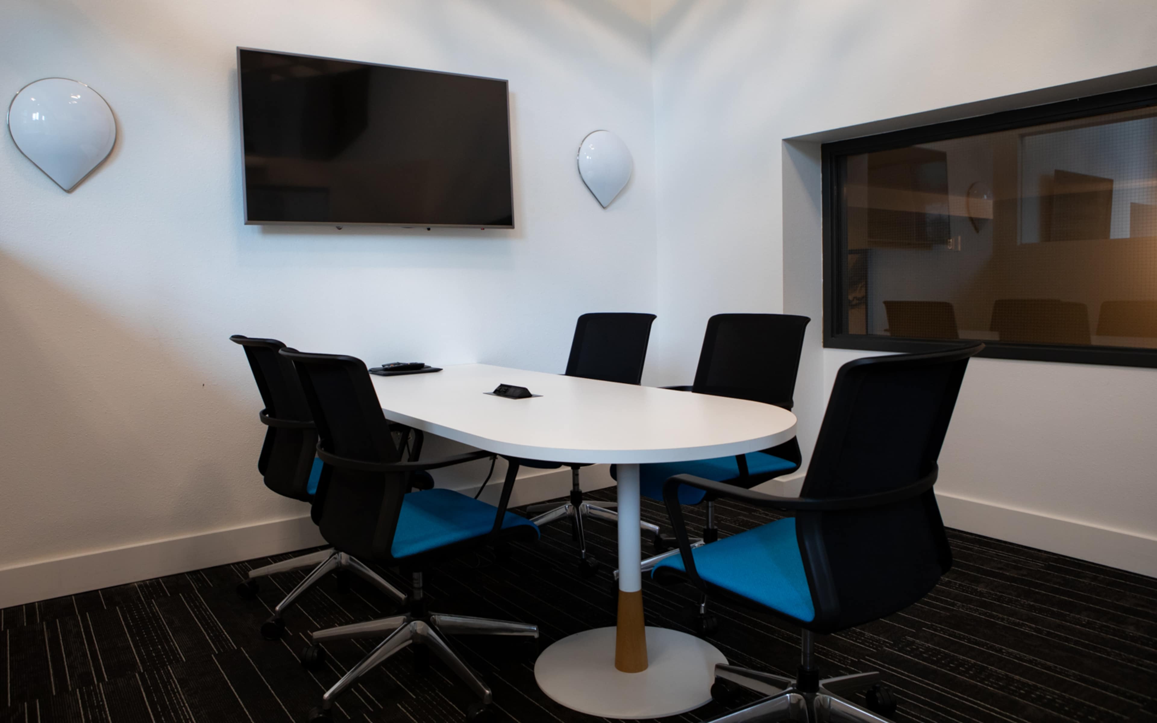 A modern conference room with a white oval table surrounded by five black chairs, a wall-mounted television, and a window.