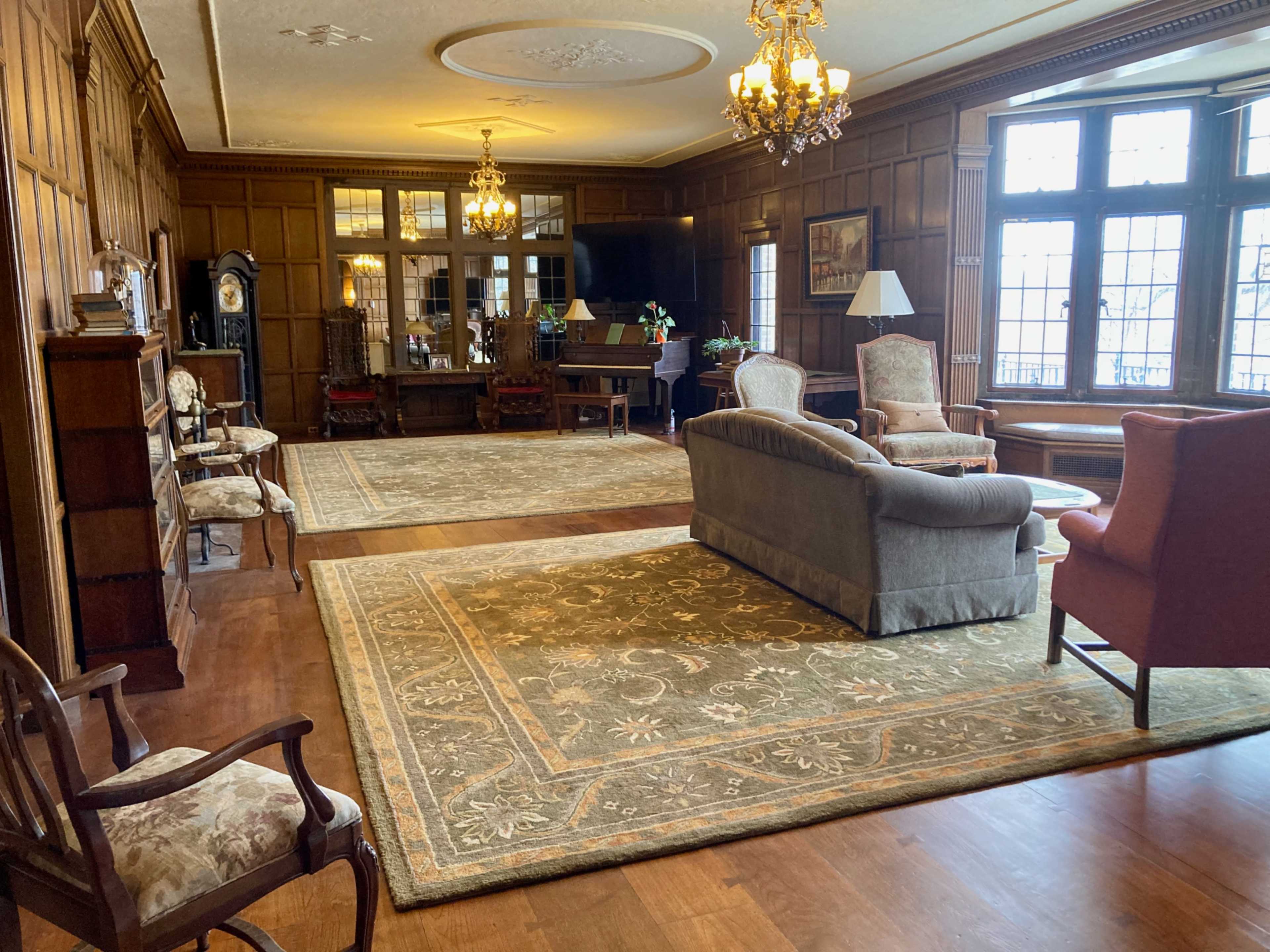 The image shows a spacious, elegantly furnished room with wooden paneling, a large area rug, and multiple light fixtures.