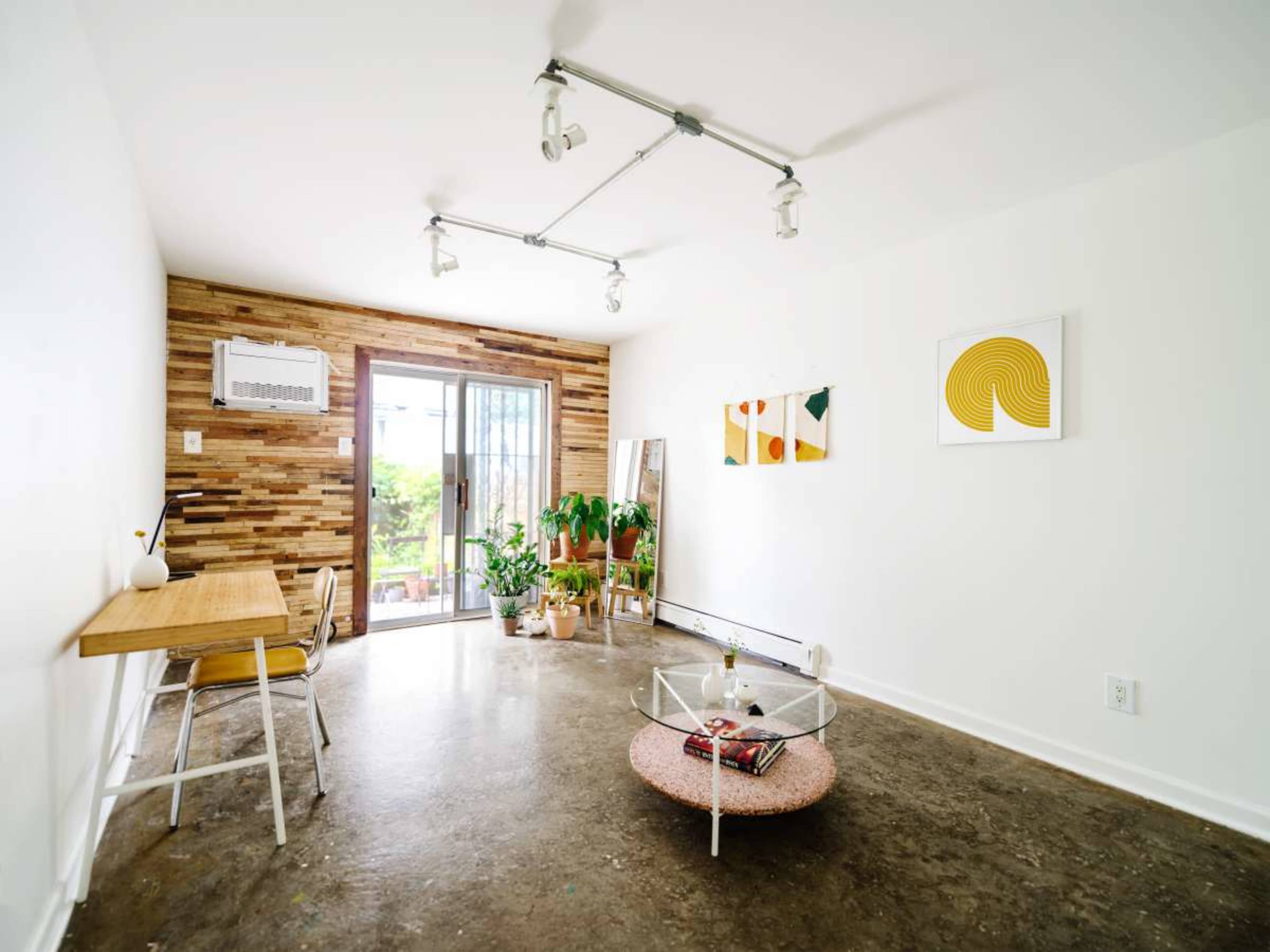 The image shows a bright, minimalist living space with a wooden accent wall, a small dining table, and a circular coffee table, along with a glass door opening to a balcony.