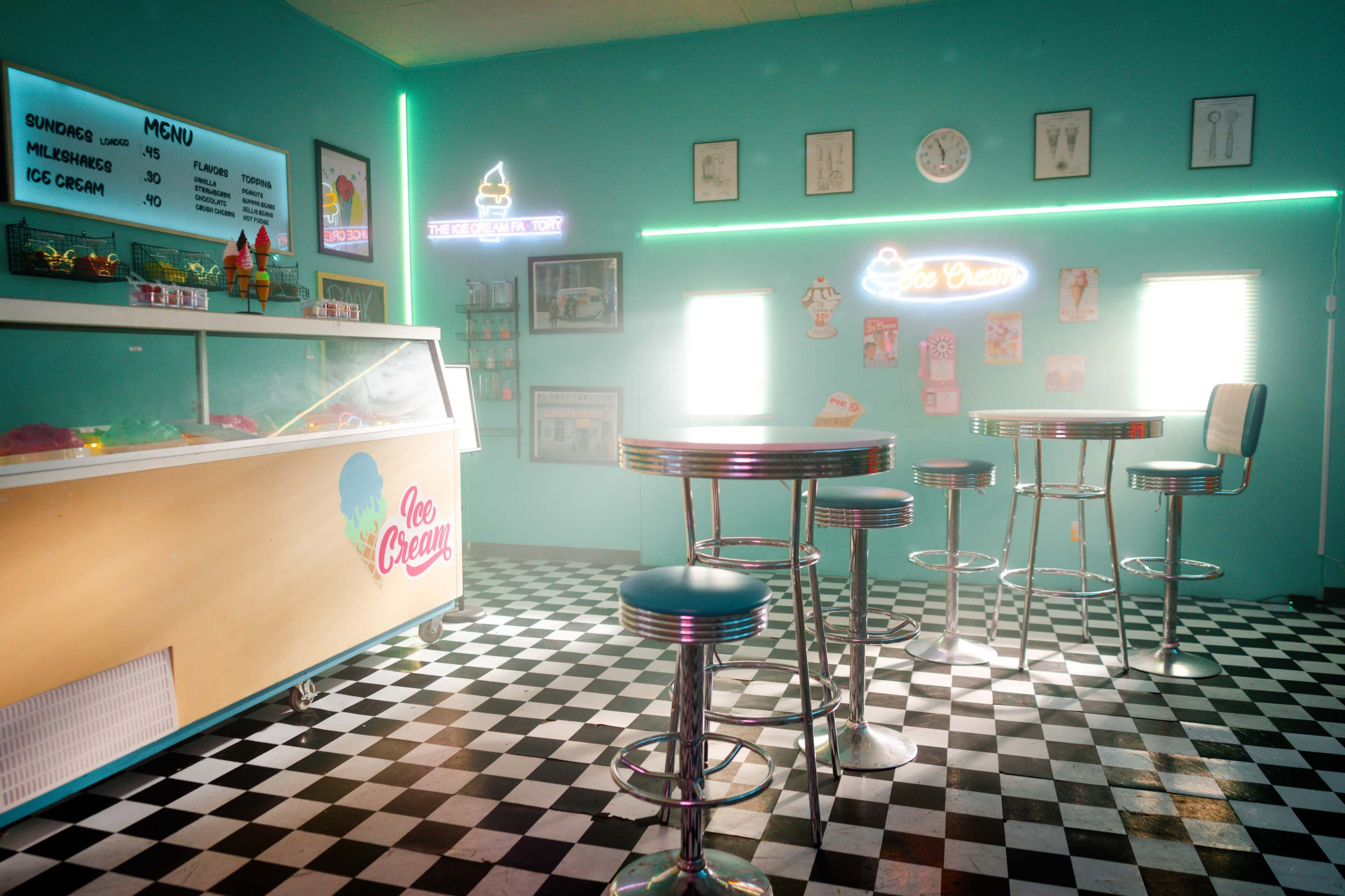The scene depicts a retro ice cream parlor with a checkered floor, a counter displaying ice cream flavors, and metal stools arranged around small tables.