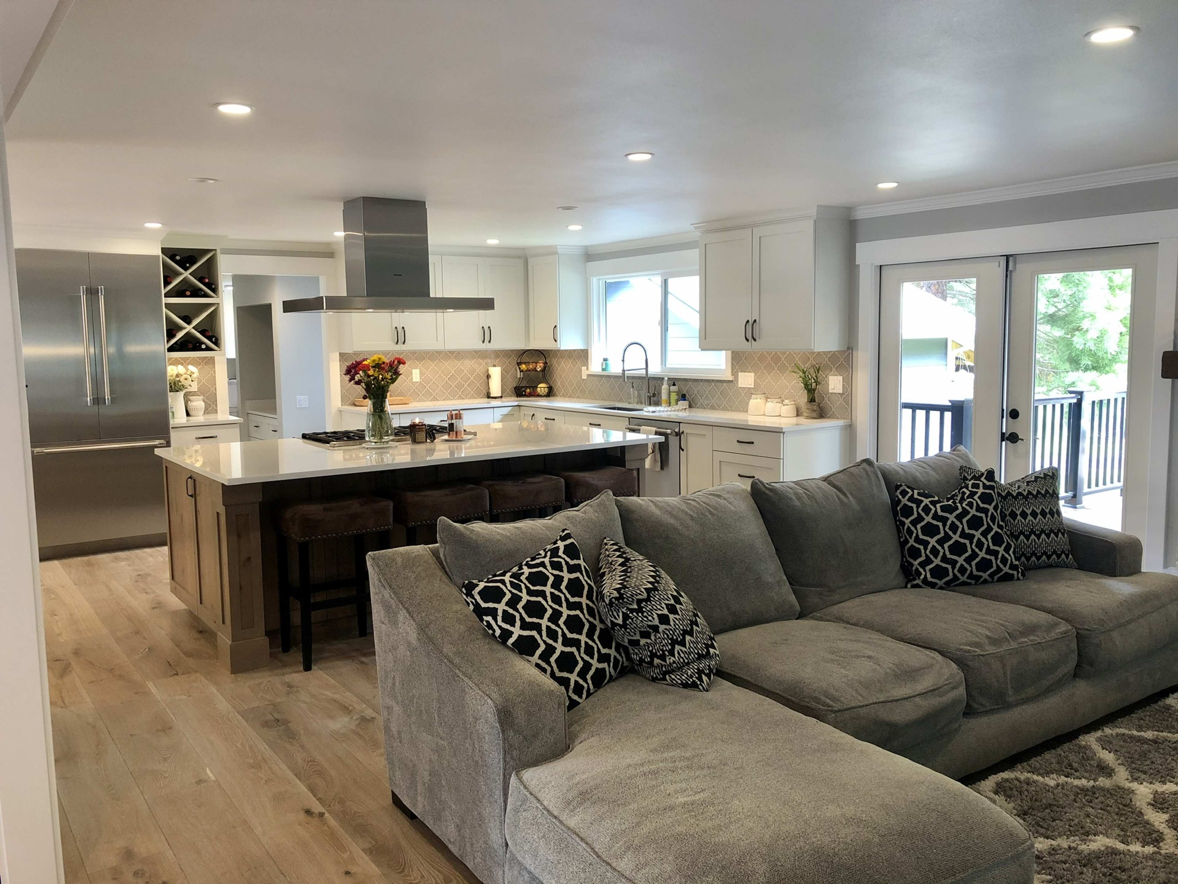 The image shows a modern open-concept kitchen and living area with a gray sectional sofa facing a large island with bar stools and stainless steel appliances.