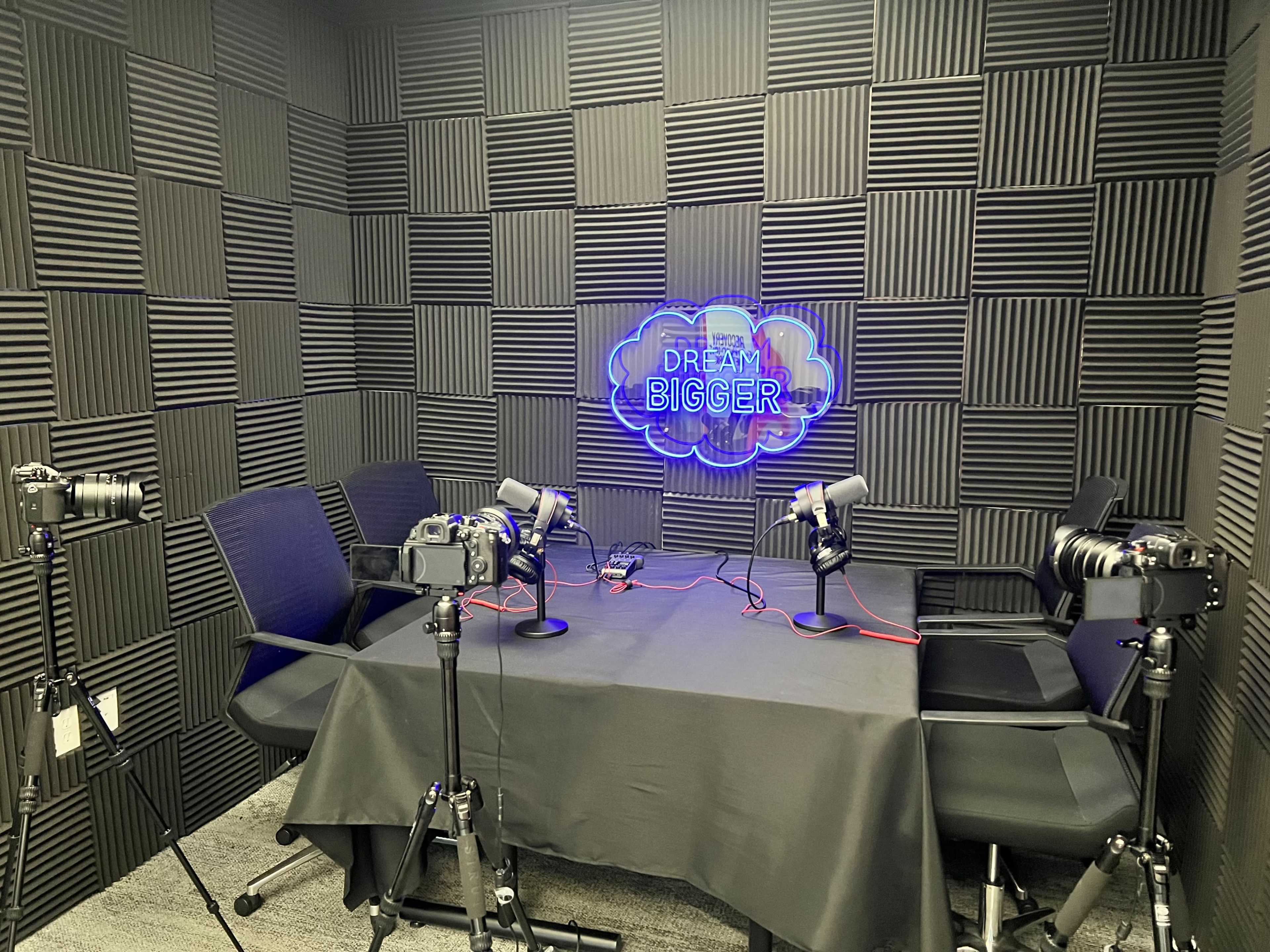 A podcasting studio featuring two microphones, two cameras, and a neon sign on a textured wall.