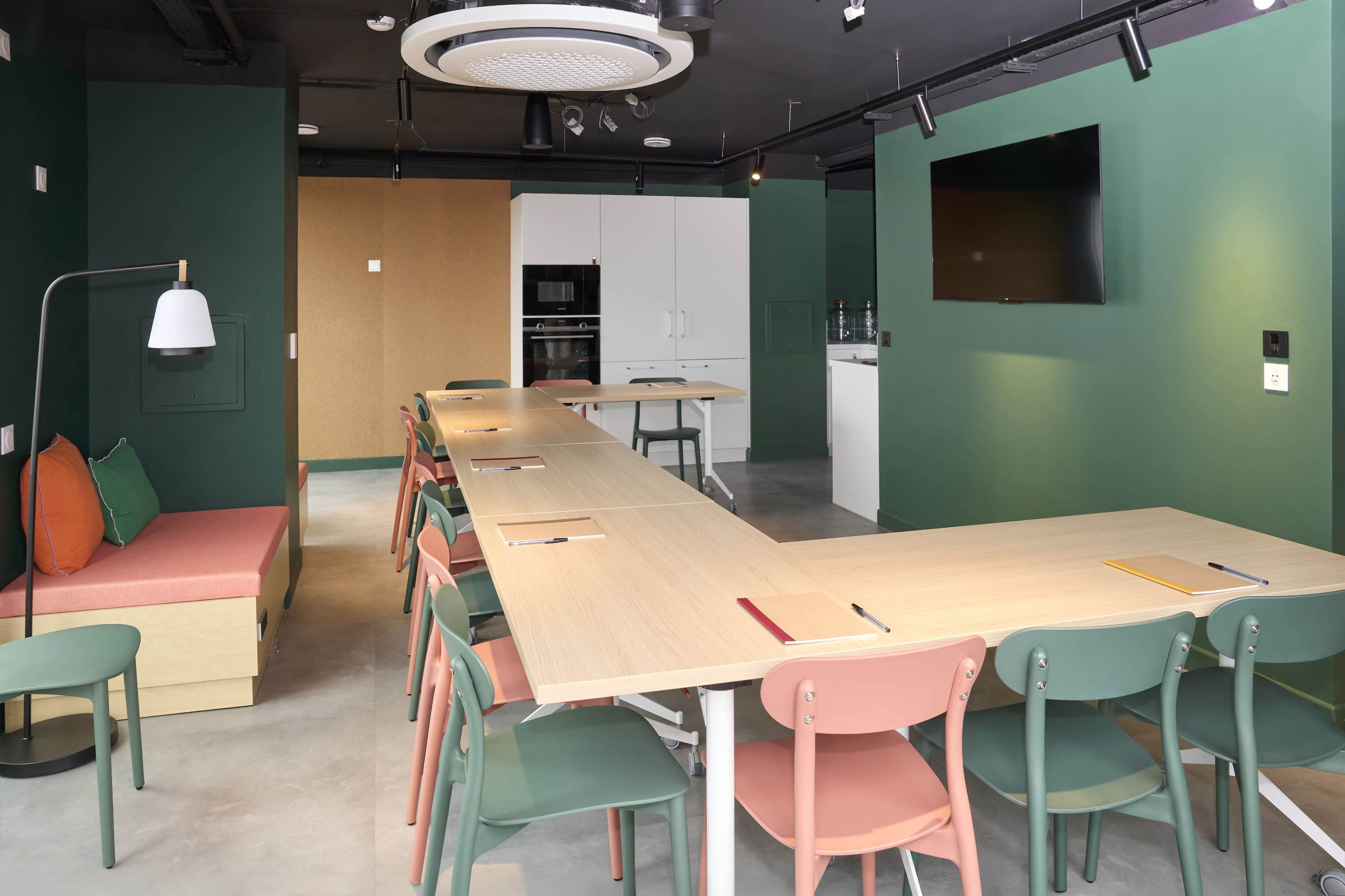 The image shows a modern conference room with a long table, colorful chairs, and a casual seating area, featuring green walls and a mounted screen.