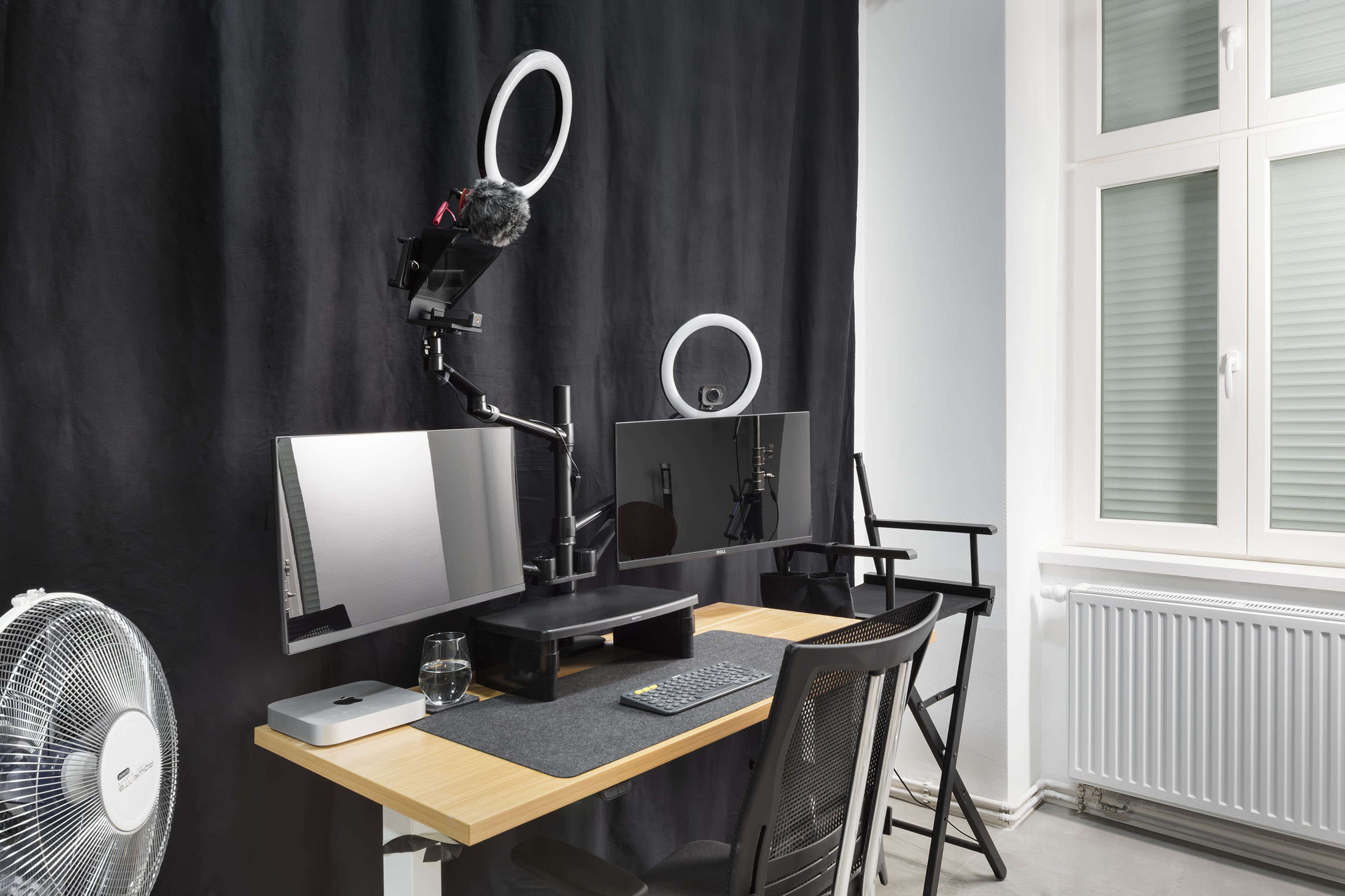 A modern workspace with two computer monitors, a microphone on a stand, a ring light, a fan, and a glass of water on a desk.