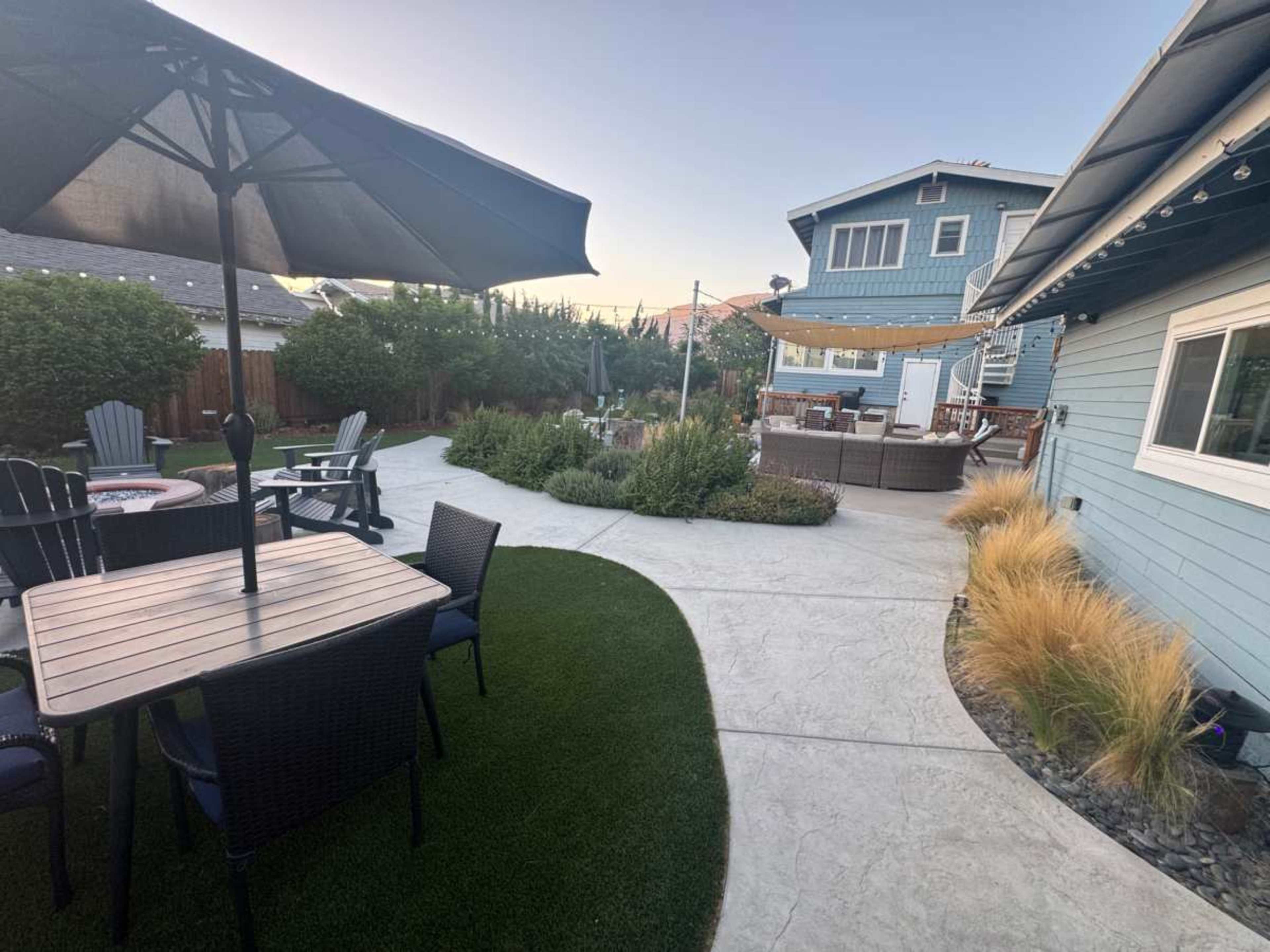 A landscaped backyard features a patio with seating, an umbrella, and a pathway leading to a larger gathering area.