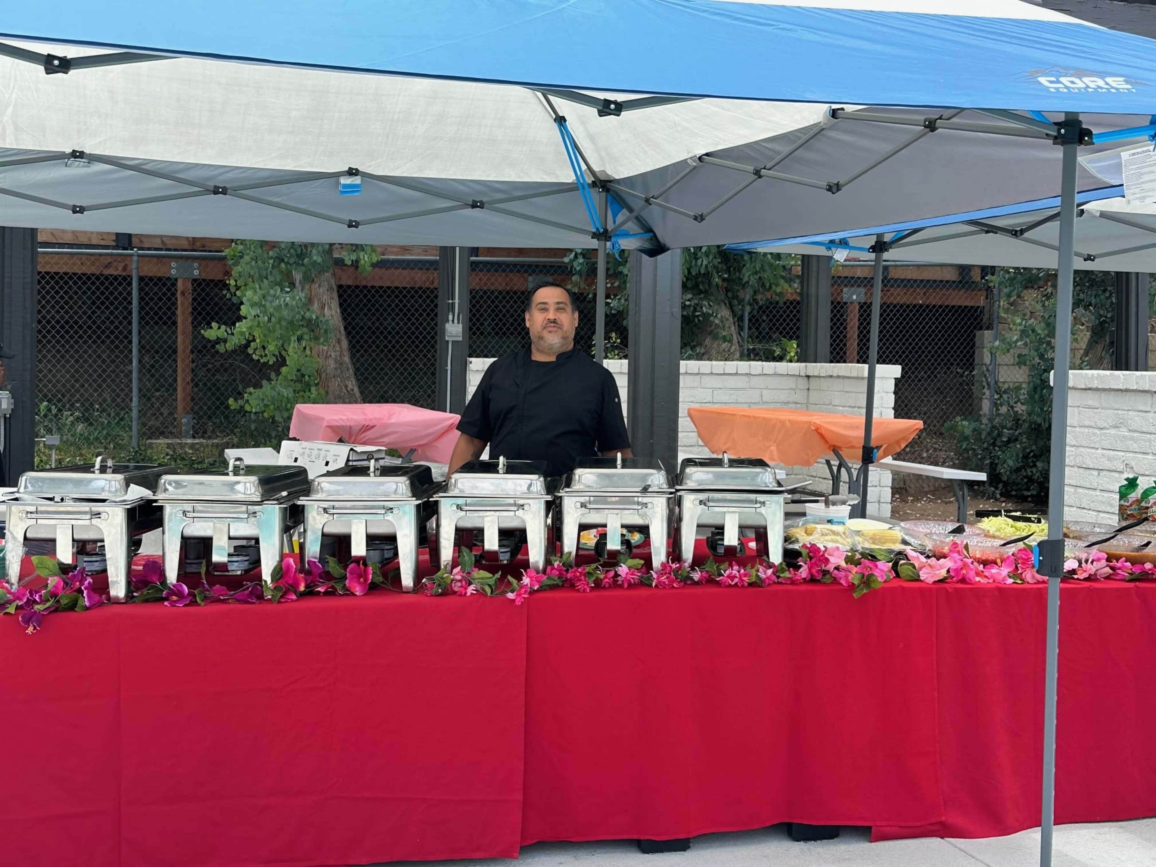 A man stands behind a table covered with a red cloth, showcasing several food trays under a tent at an outdoor event.