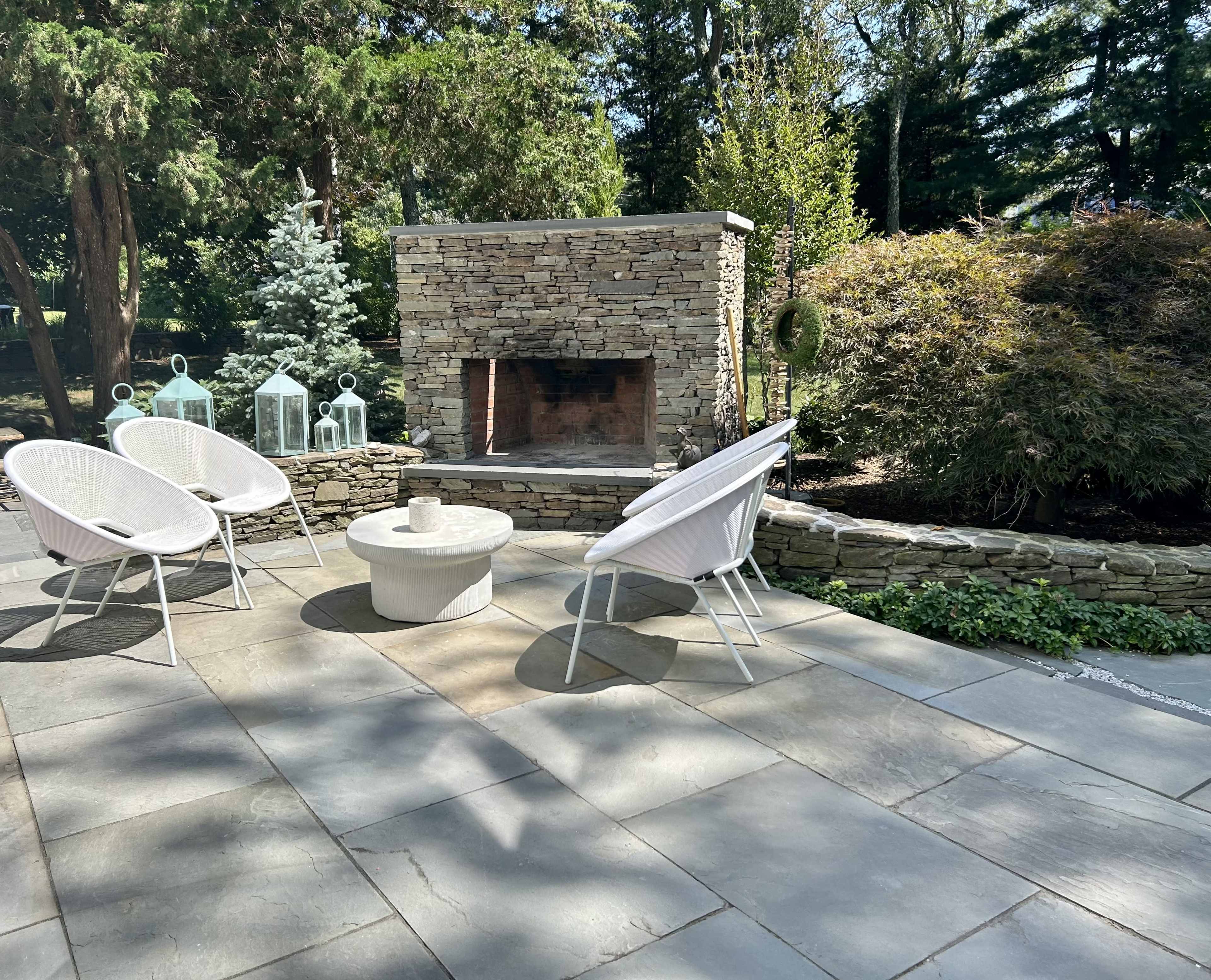 A stone patio area with three whitechairs arranged around a circular table, complemented by a stone fireplace and lush greenery in the background.