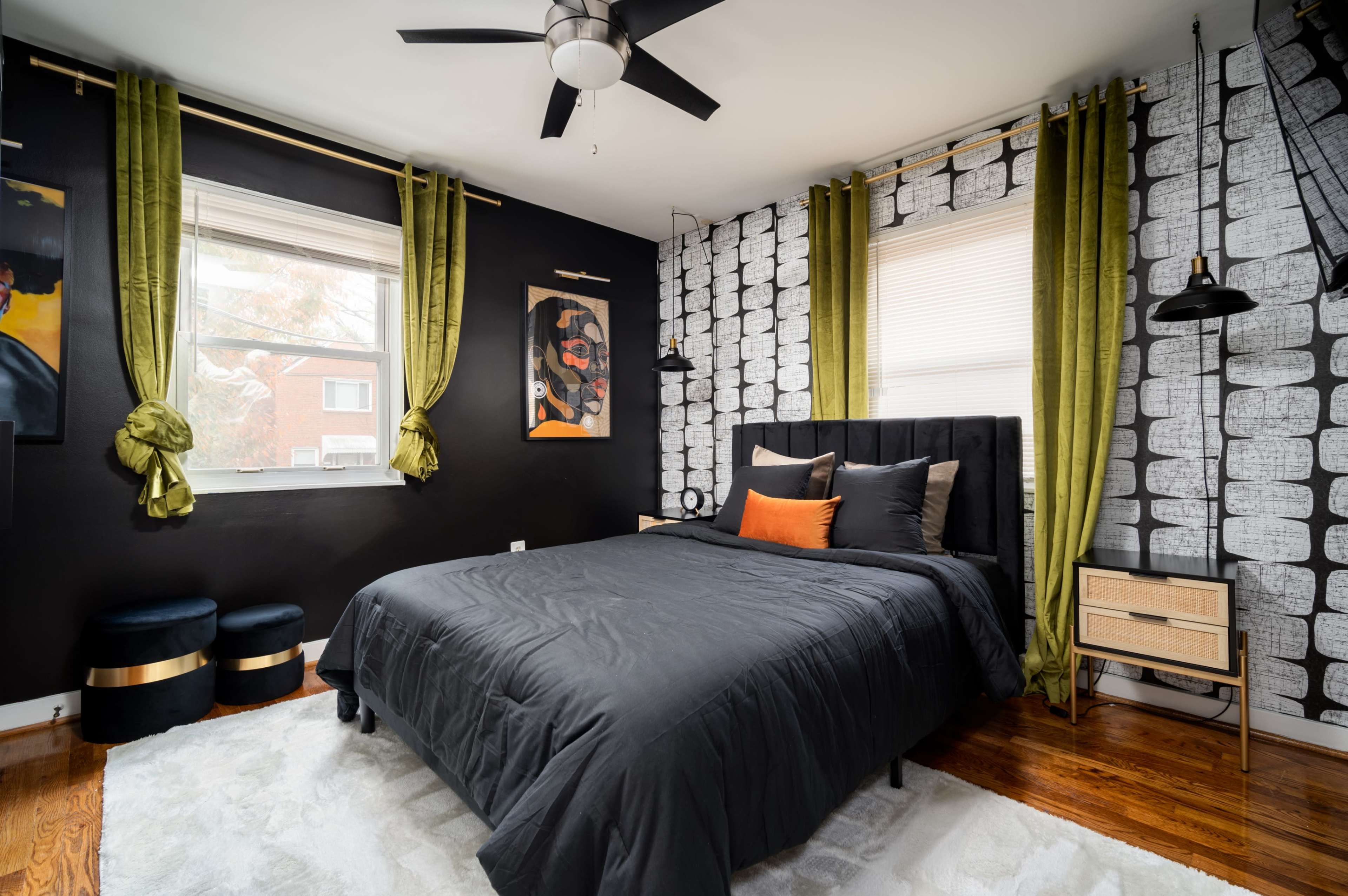 The room features a black bed with gray and orange accent pillows, flanked by green curtains, against a backdrop of patterned wallpaper and wooden flooring.