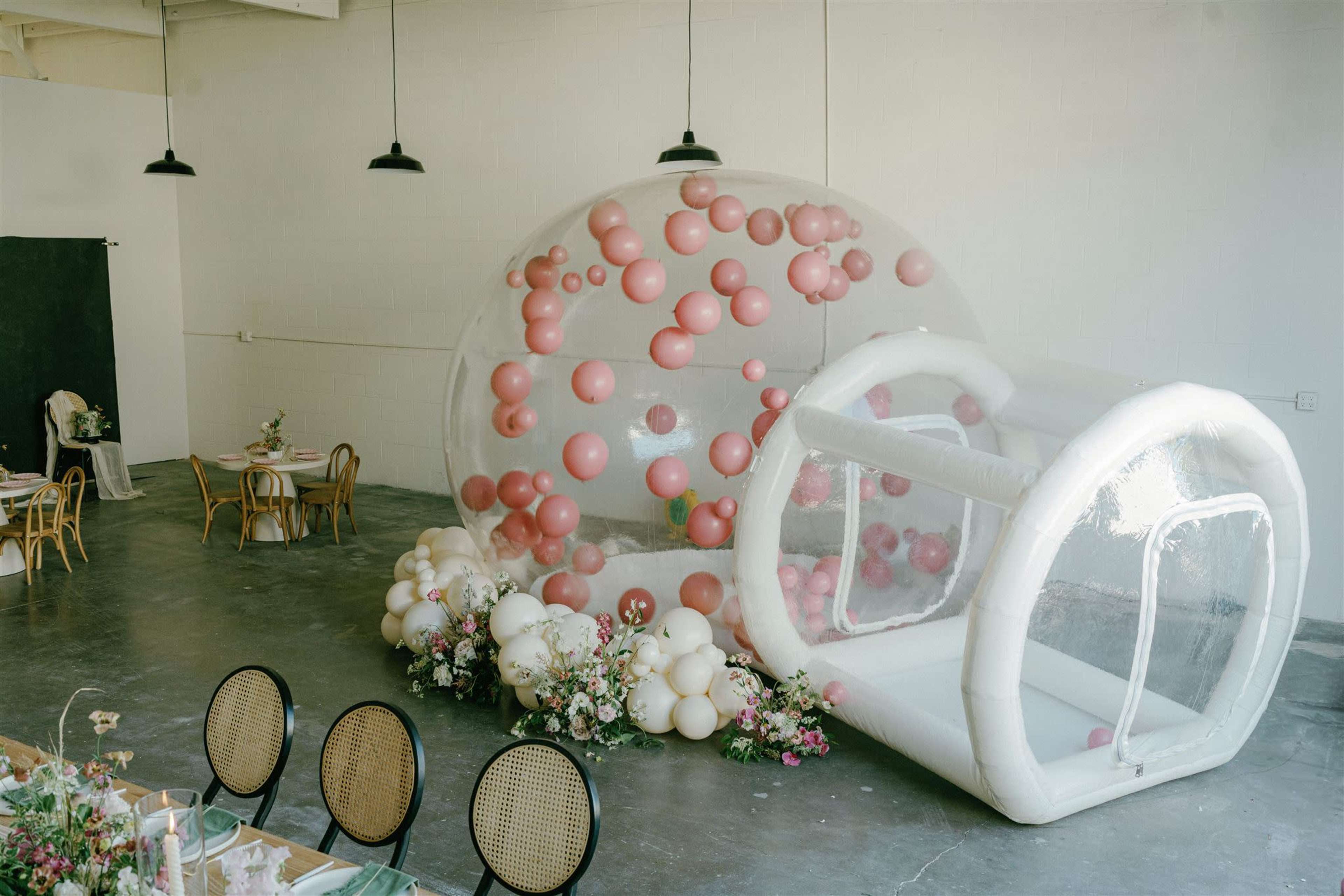 A spacious indoor venue featuring a large clear inflatable dome filled with colorful balloons and a smaller inflatable structure beside it, surrounded by floral arrangements and seating areas.