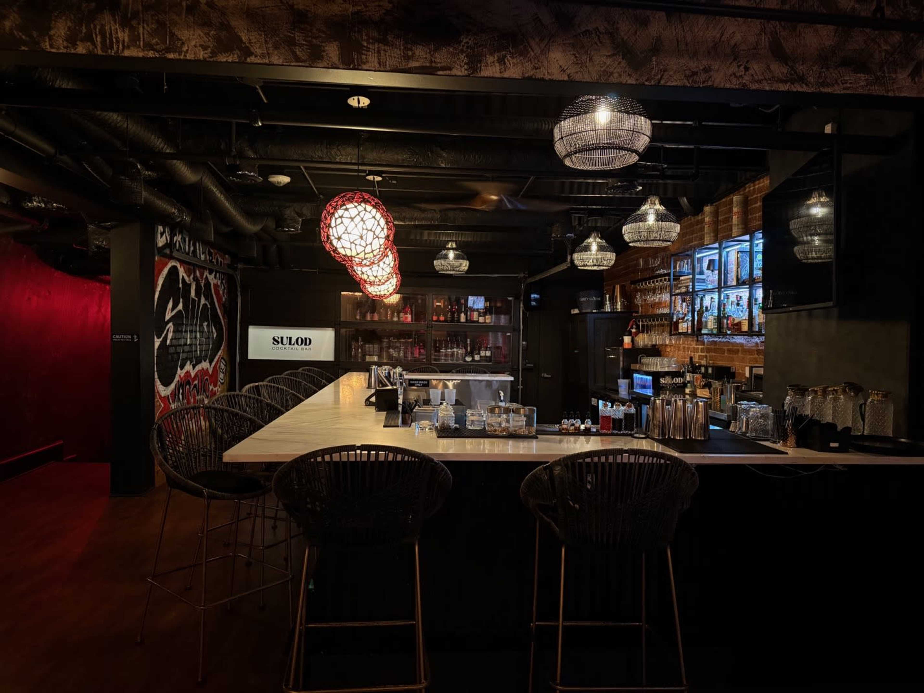 The image shows a dimly lit bar with a long counter, high-backed stools, and decorative pendant lights hanging from the ceiling.