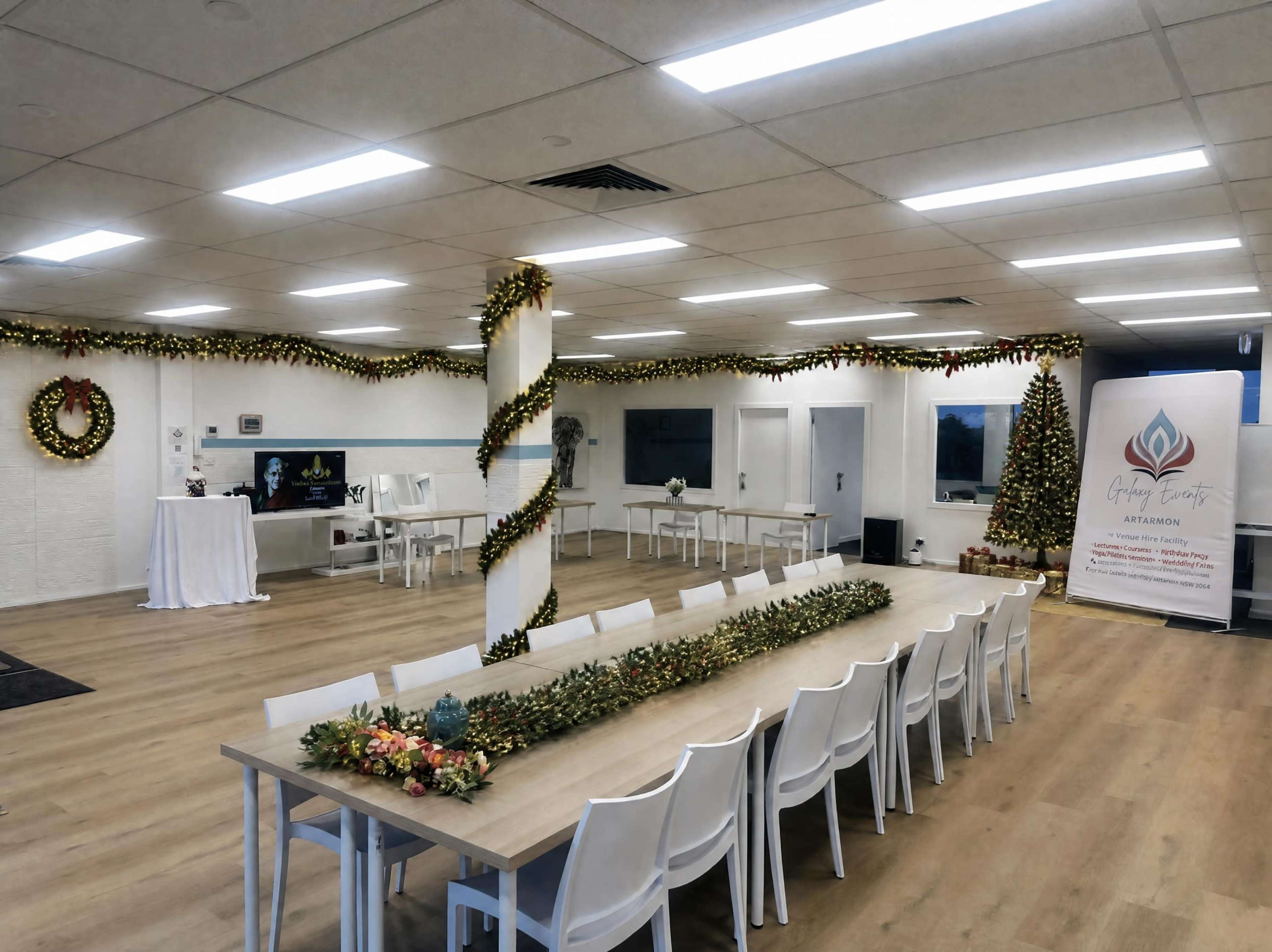 The image shows a spacious event room decorated with Christmas garlands, a large table with white chairs, and a decorated tree in the corner.