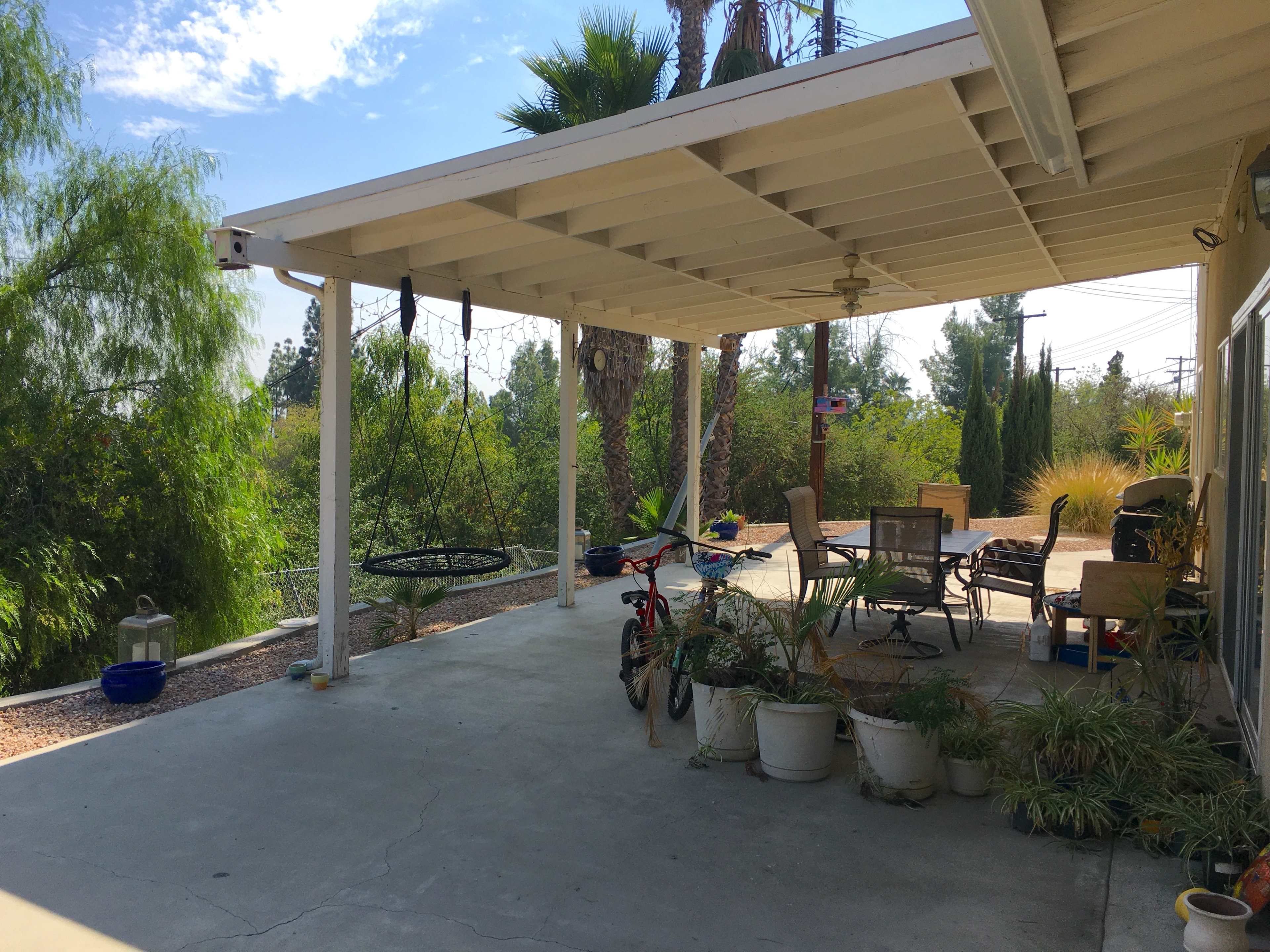 A covered patio area with potted plants, a bicycle, and outdoor furniture overlooking a green landscape.