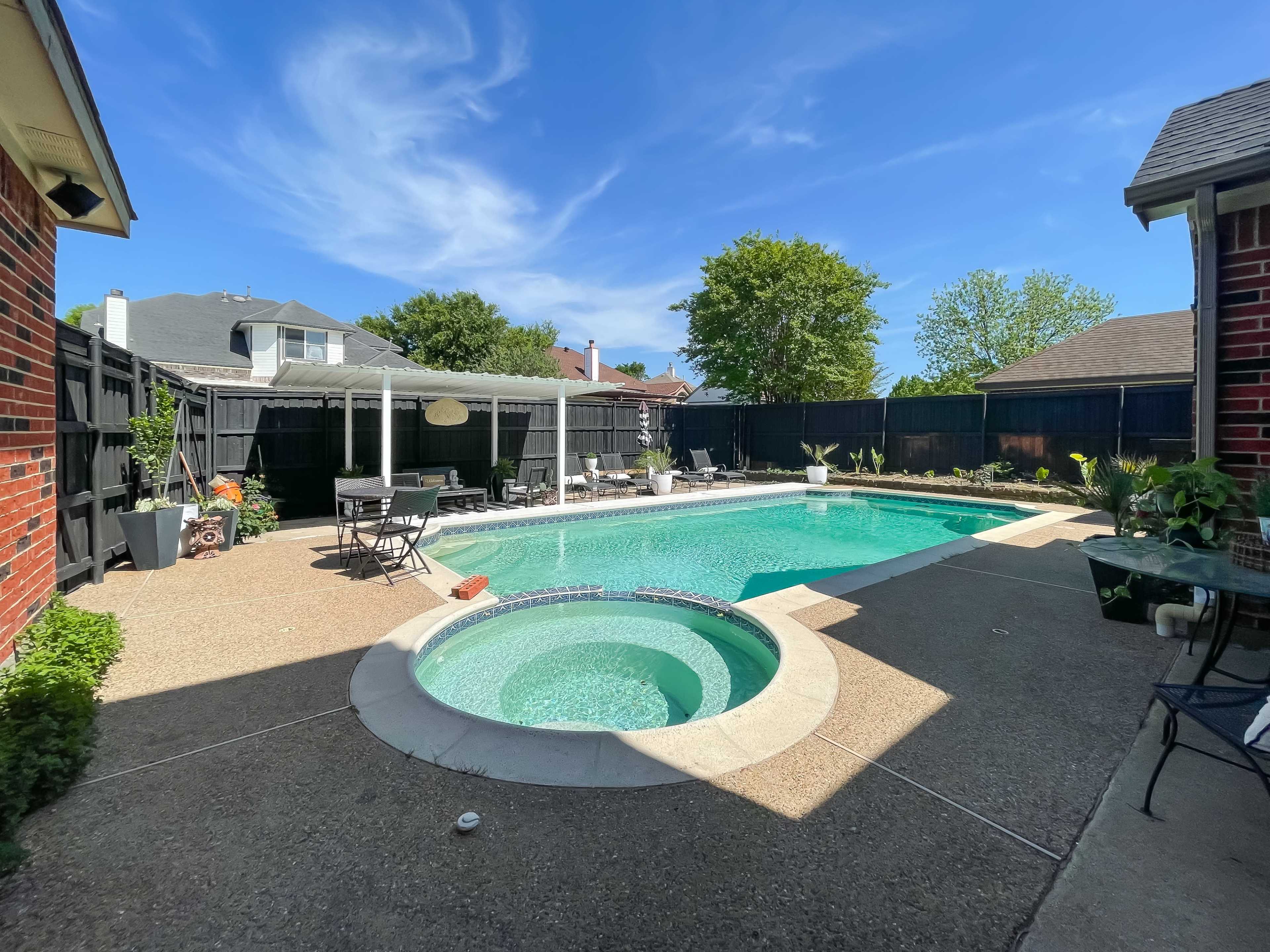 The image shows a backyard swimming pool with a built-in hot tub, surrounded by a patio area and greenery under a clear blue sky.
