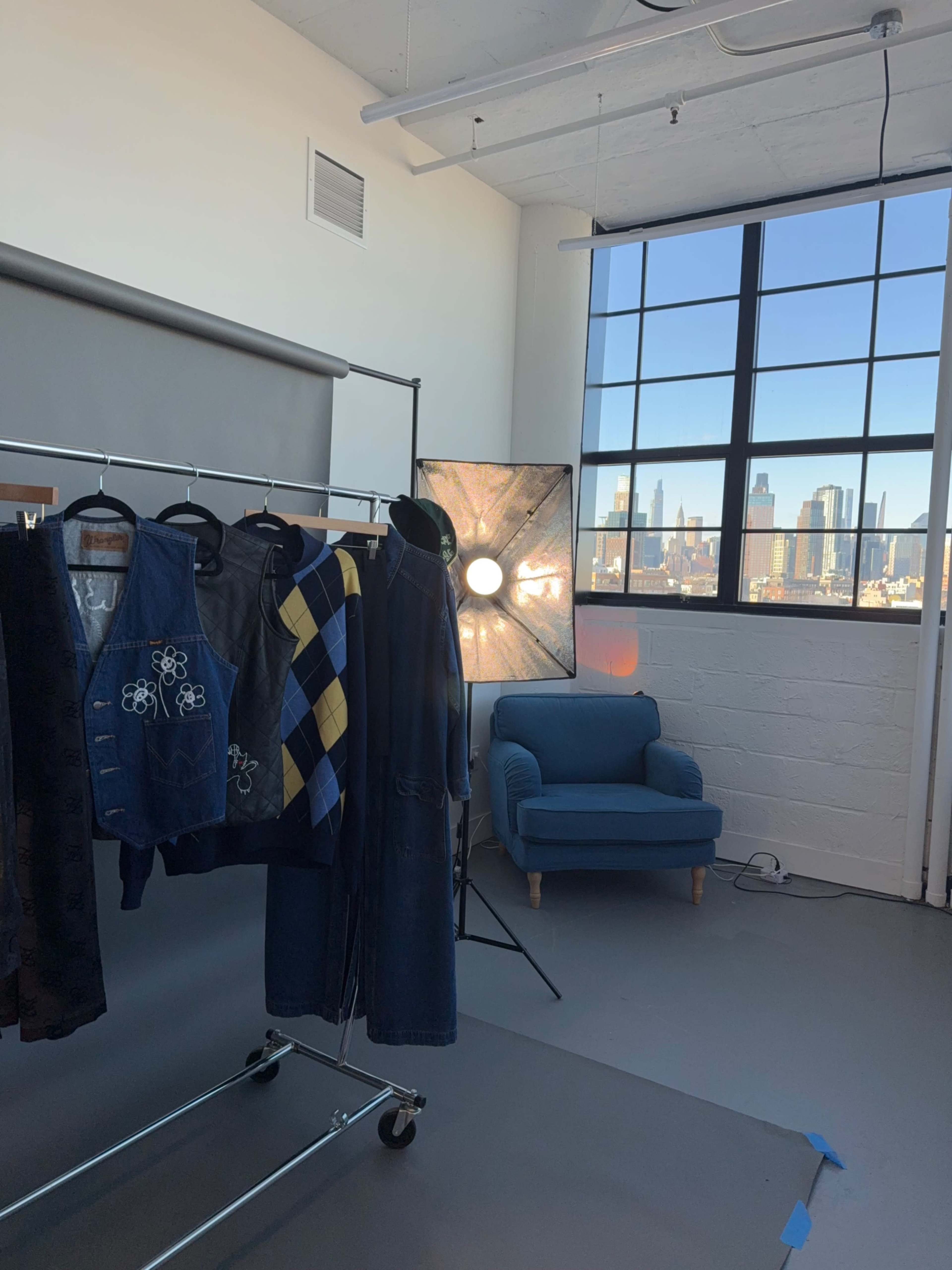 The image shows a well-lit studio setting with a clothing rack displaying various garments, a blue sofa in the corner, and large windows revealing a city skyline.