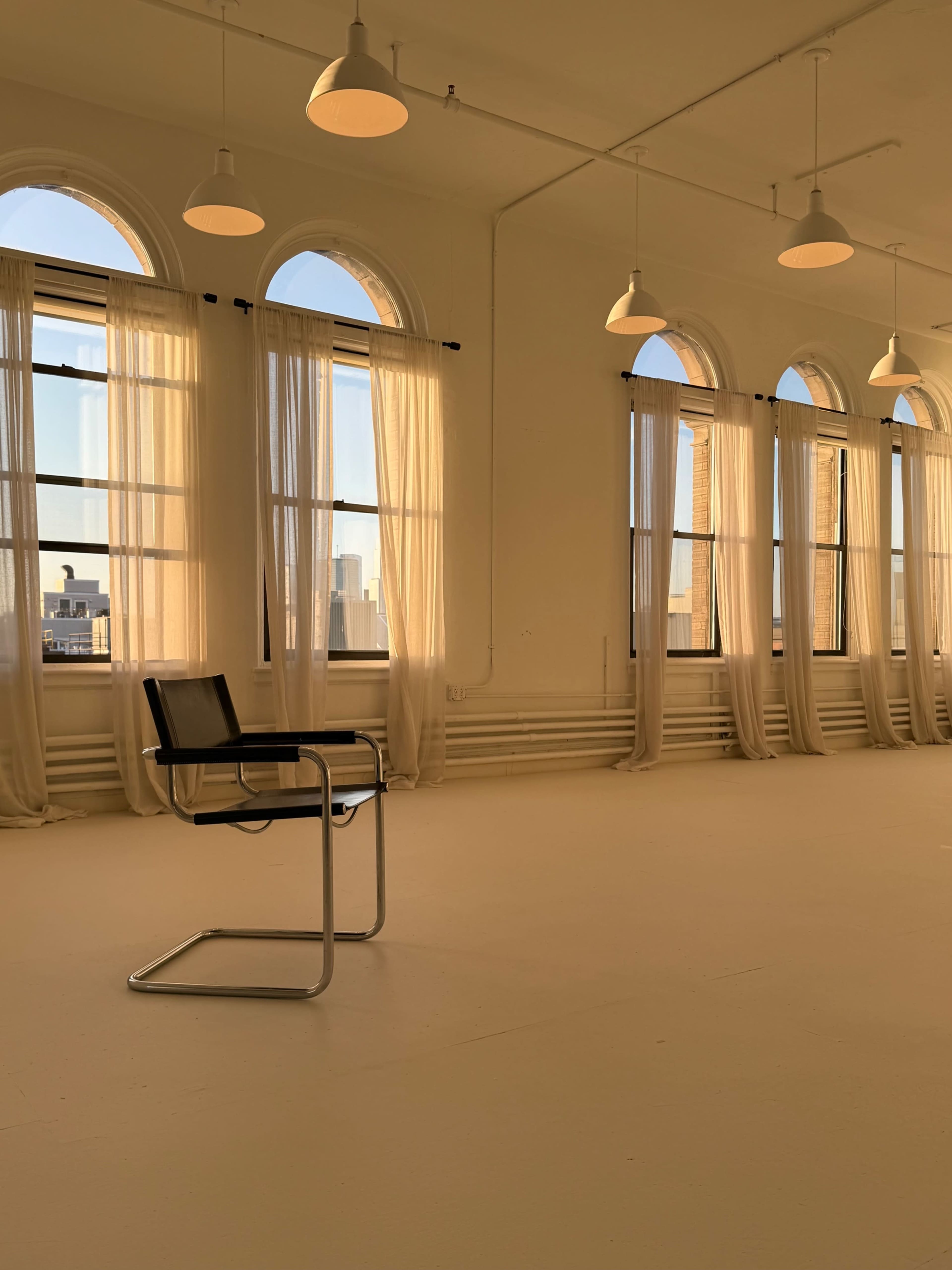 A black chair is positioned on a white floor in a brightly lit room with large arched windows and sheer curtains.