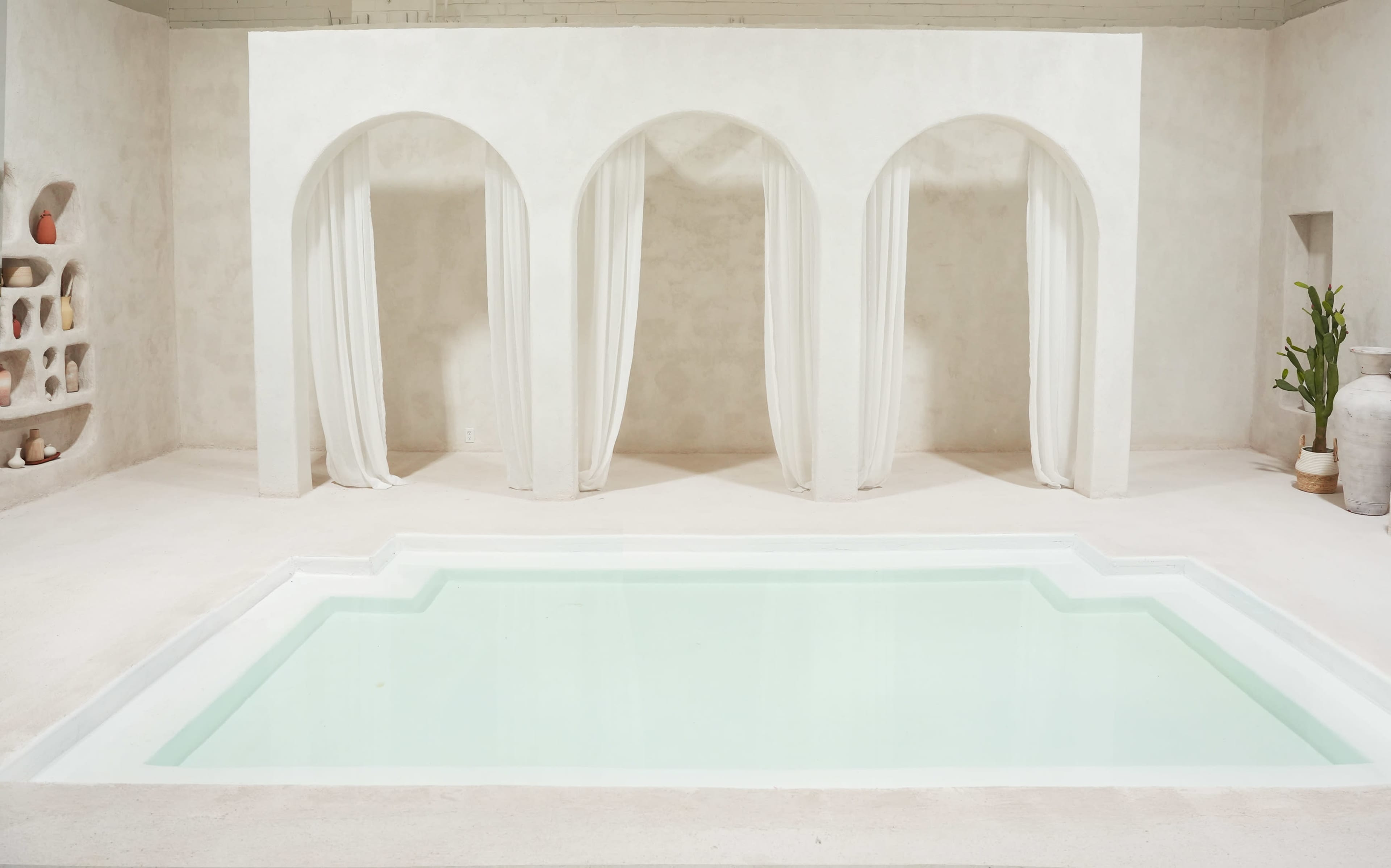 A minimalist indoor pool area features three large arched openings with white drapes and a light-colored, tiled pool.