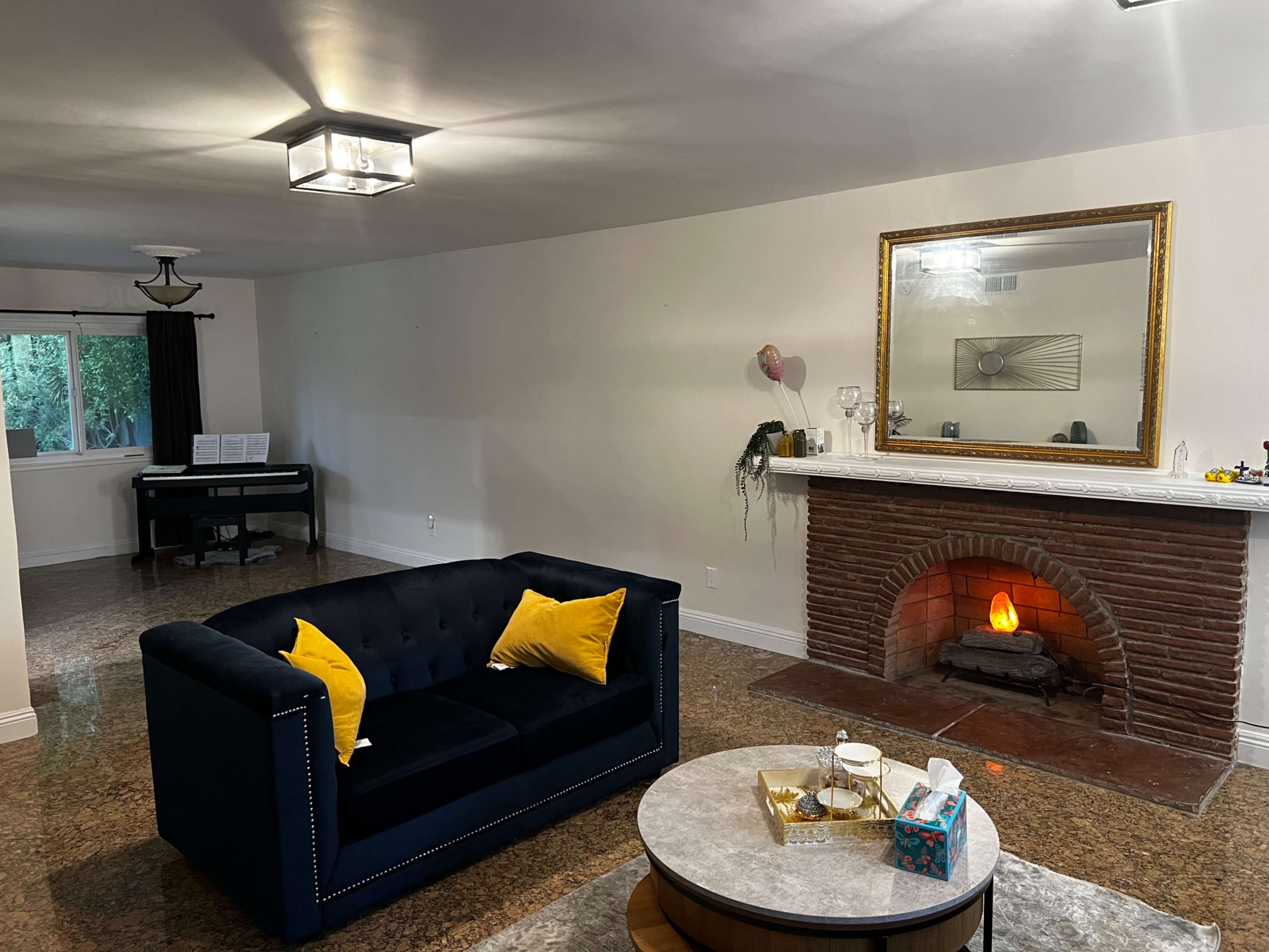A living room with a dark blue sofa, yellow cushions, a fireplace with a fire, a round coffee table, and a piano in the background.