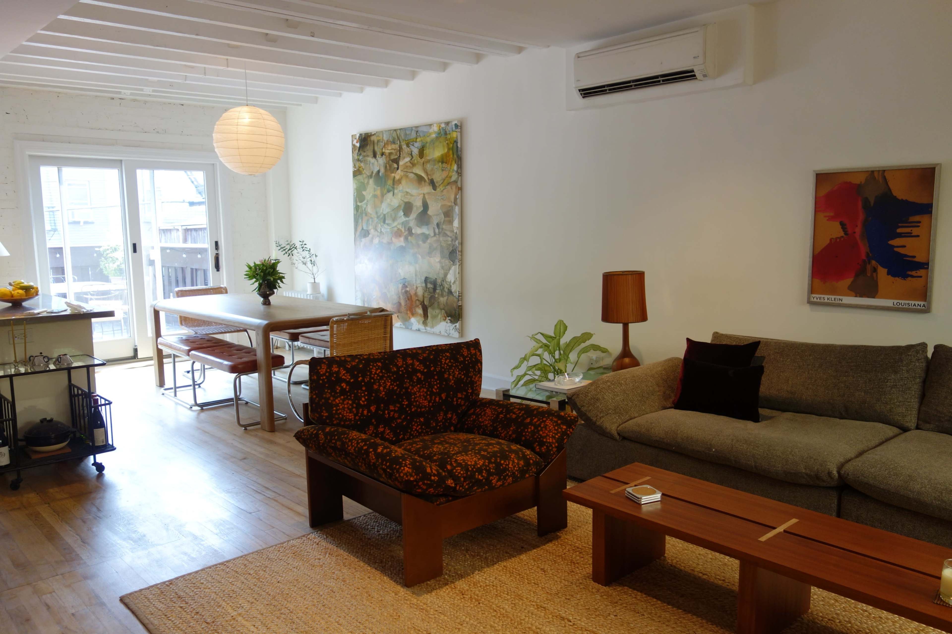 The image shows a modern living space with a brown floral-patterned armchair, a gray sofa, a wooden coffee table, a dining area with a table and chairs, and large windows leading to an outdoor space.