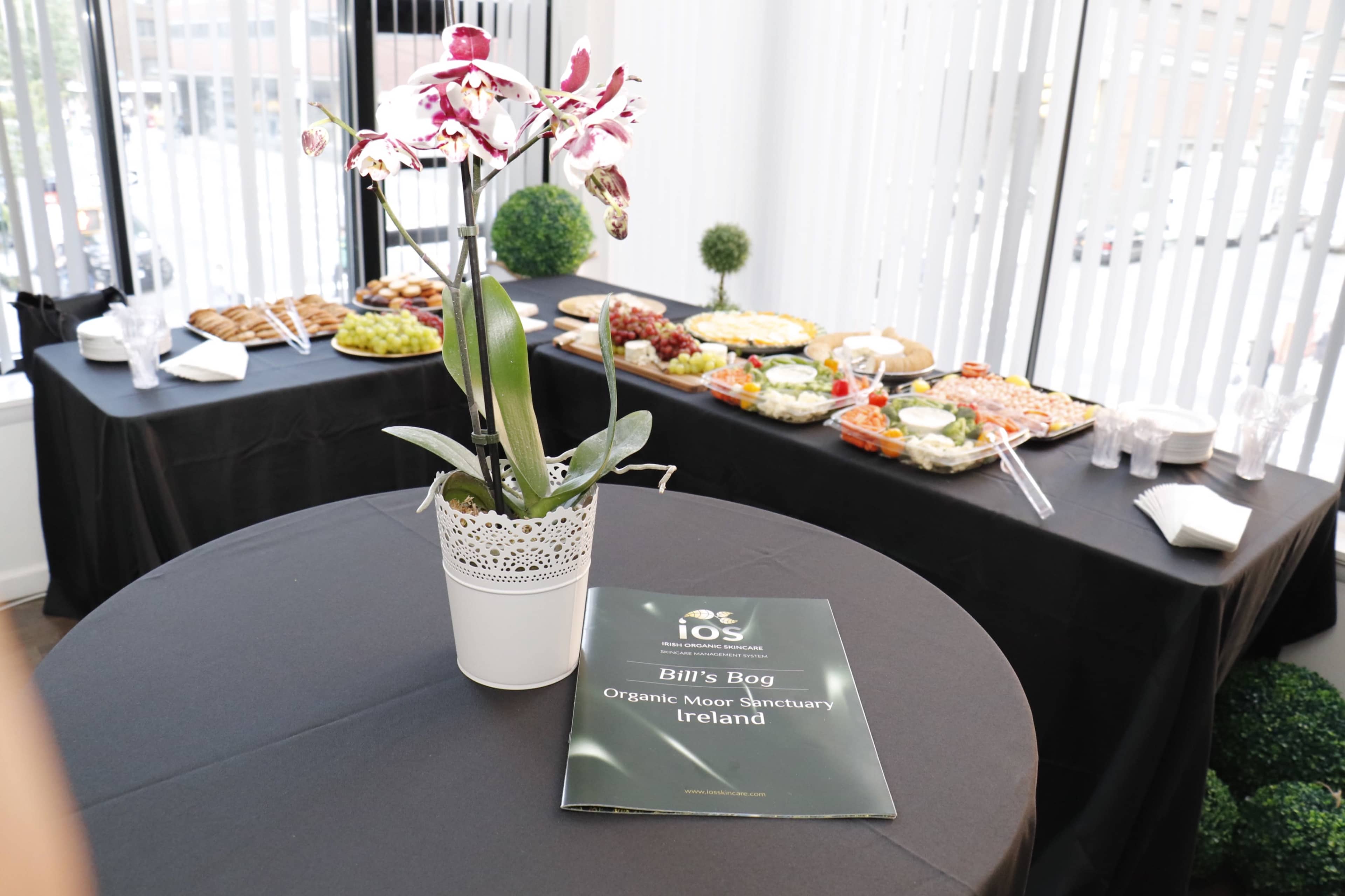 A table is set with a variety of food items, and a potted orchid is placed on a black tablecloth alongside a brochure titled "Bill's Bog Organic Moor Sanctuary Ireland."