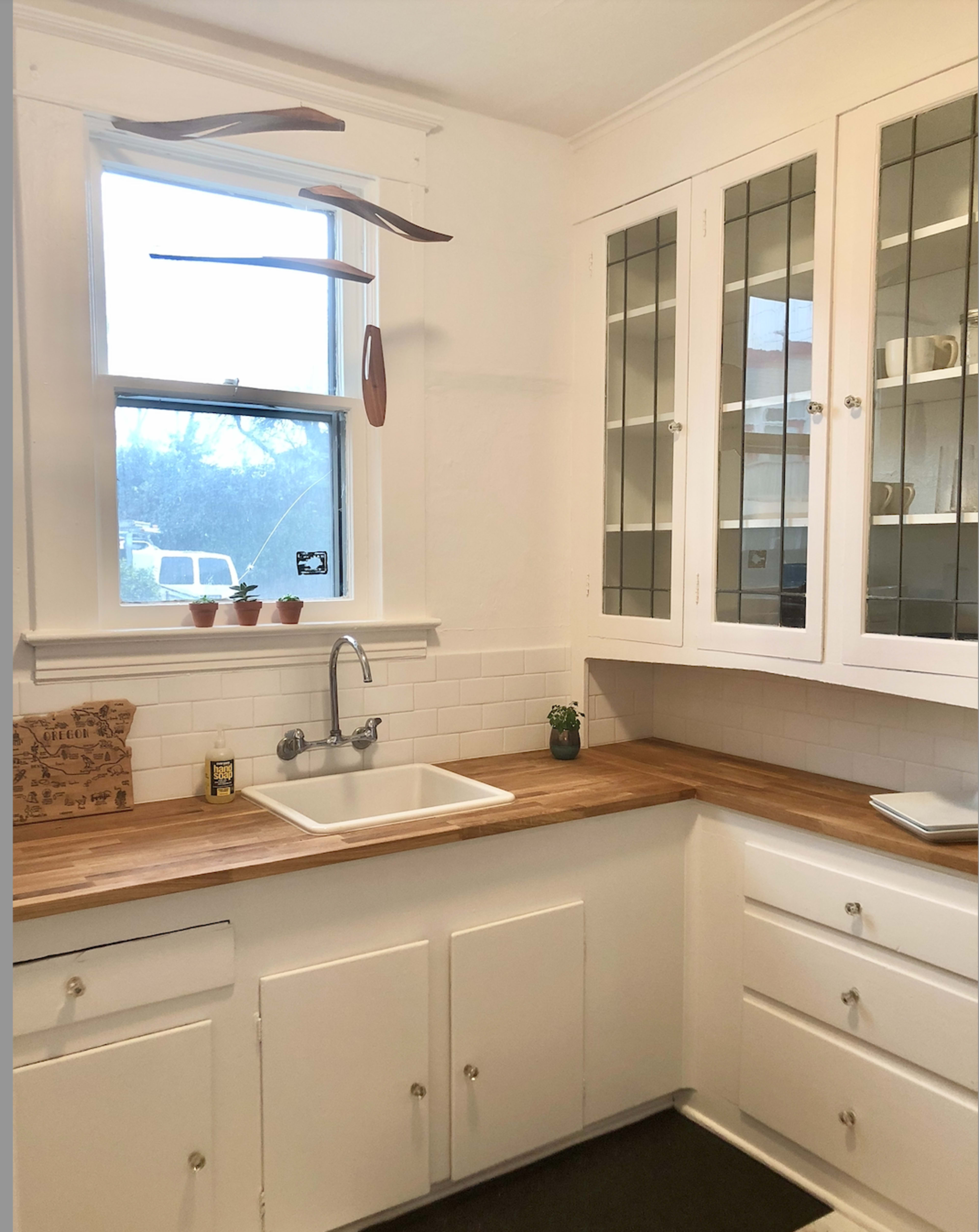 The image shows a modern kitchen with white cabinets, a wooden countertop, a farmhouse sink, and a window letting in natural light.