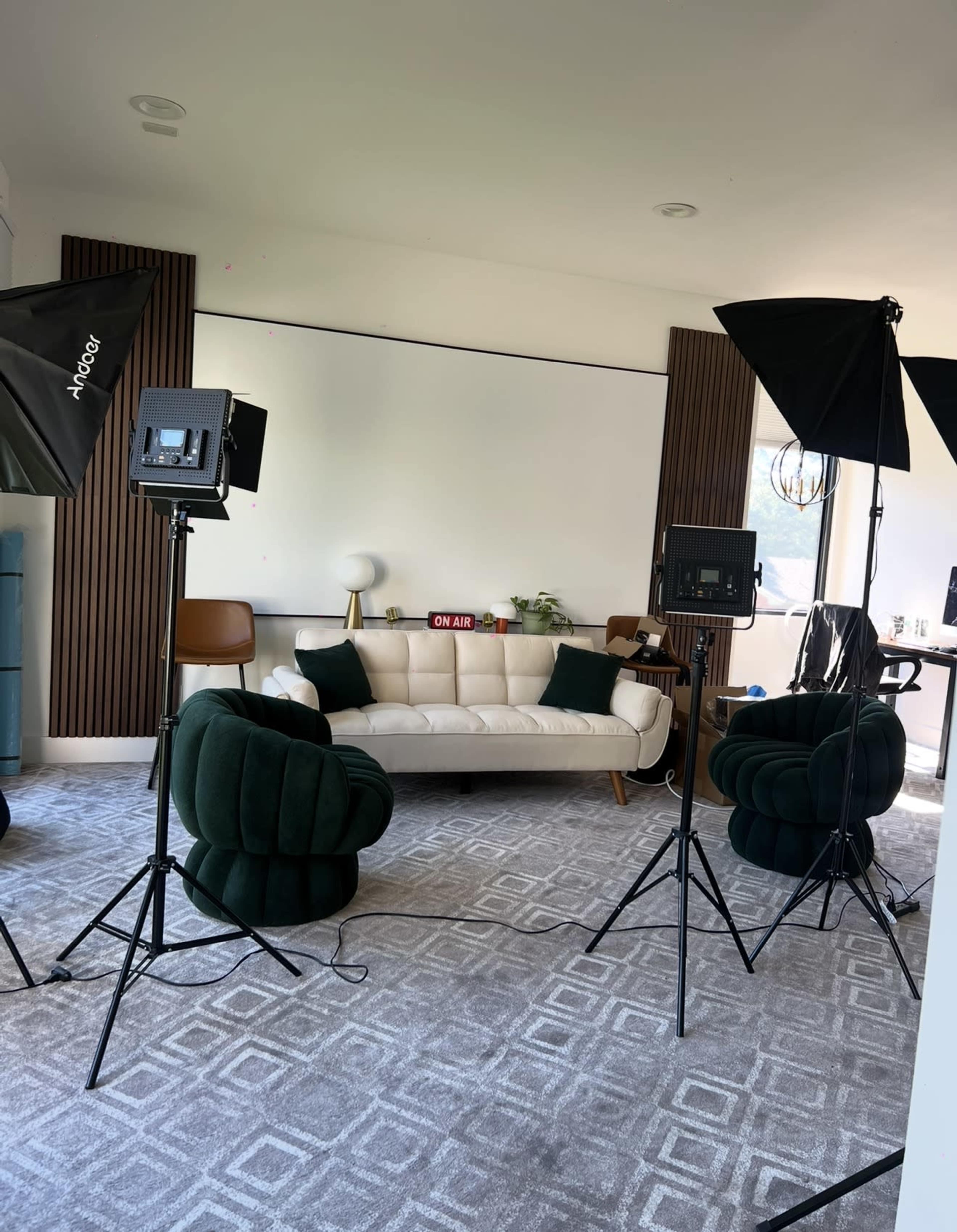 The image shows a well-lit studio setup featuring a white sofa, two green chairs, and professional lighting equipment in front of a blank wall.