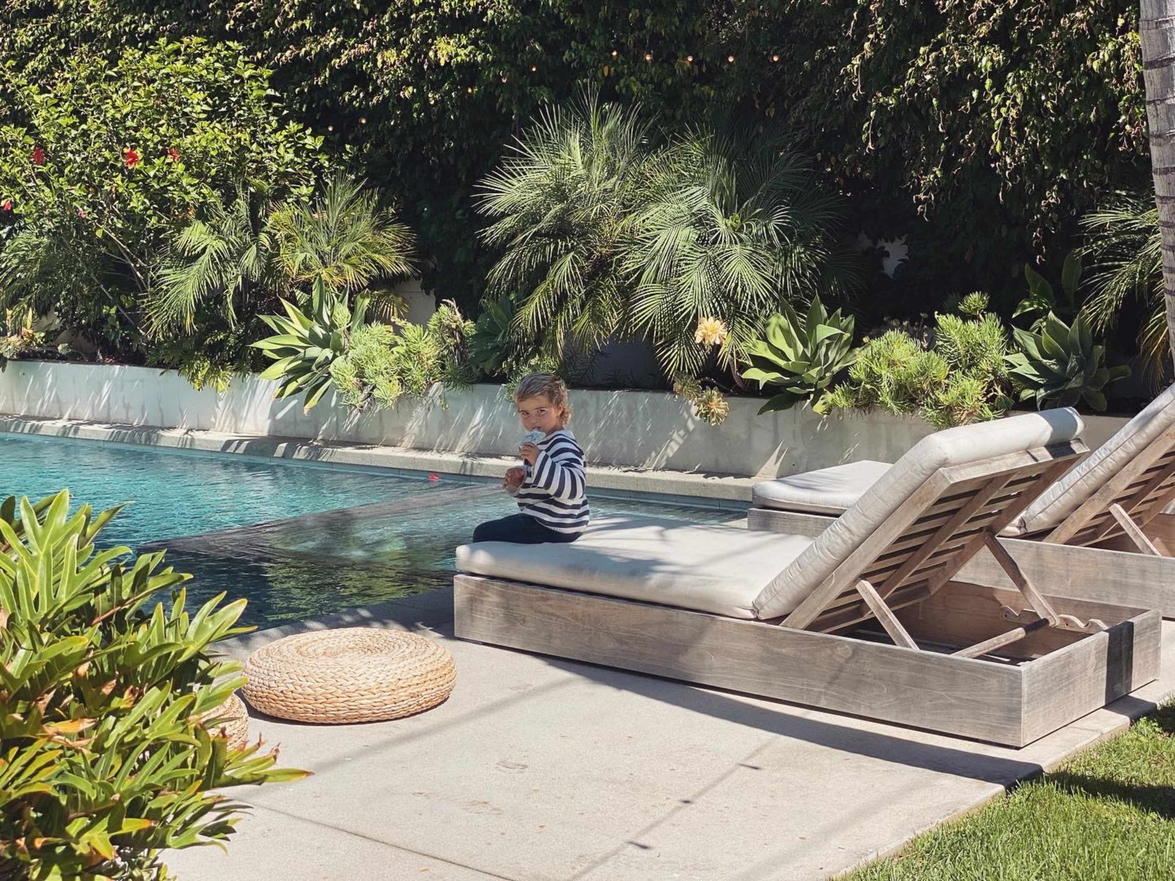 A child in a striped shirt sits on the edge of a sunbed beside a swimming pool surrounded by green plants.