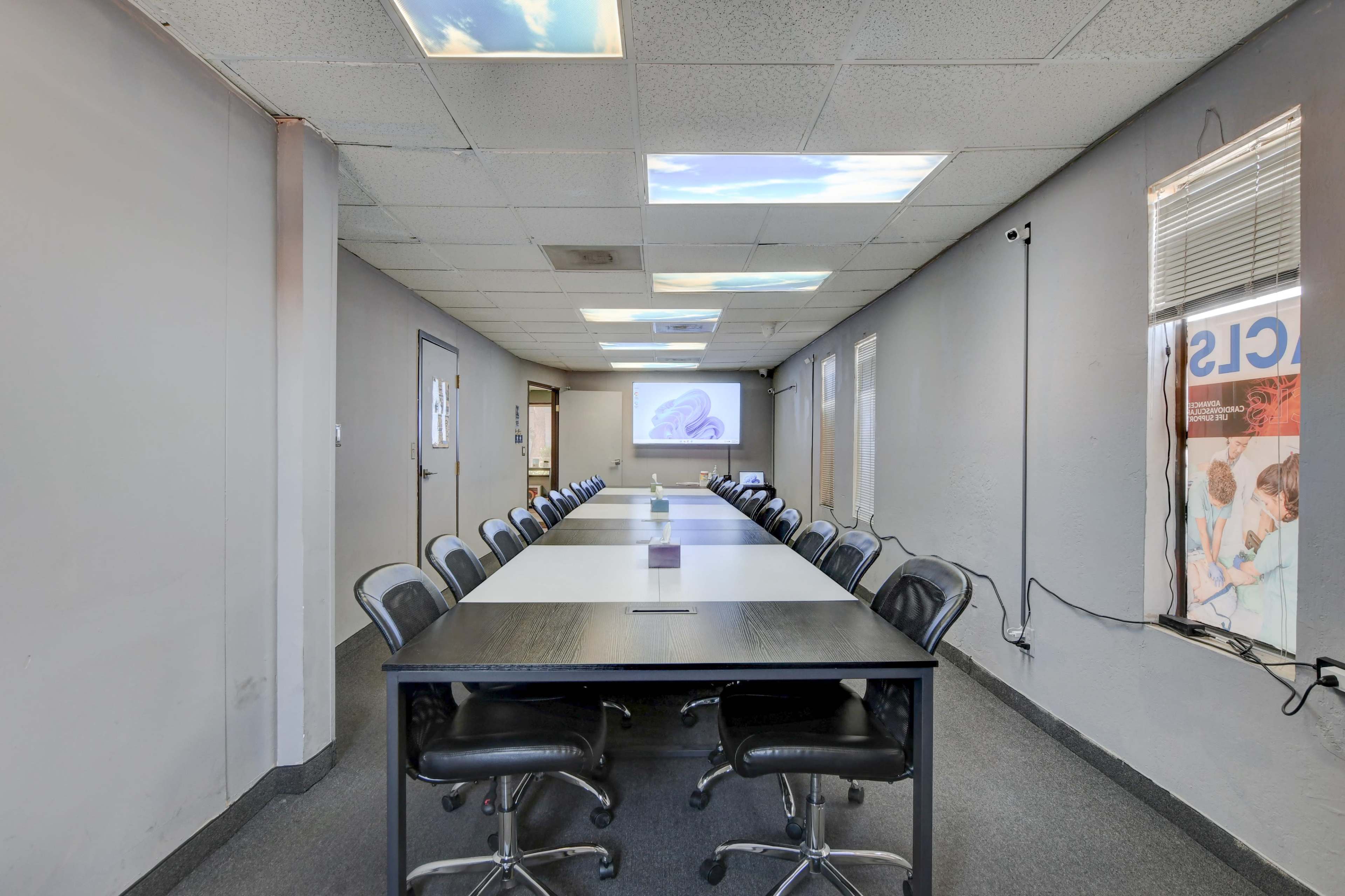 The image shows a long conference room table surrounded by chairs, with a projector screen at one end and large windows on the ceiling.