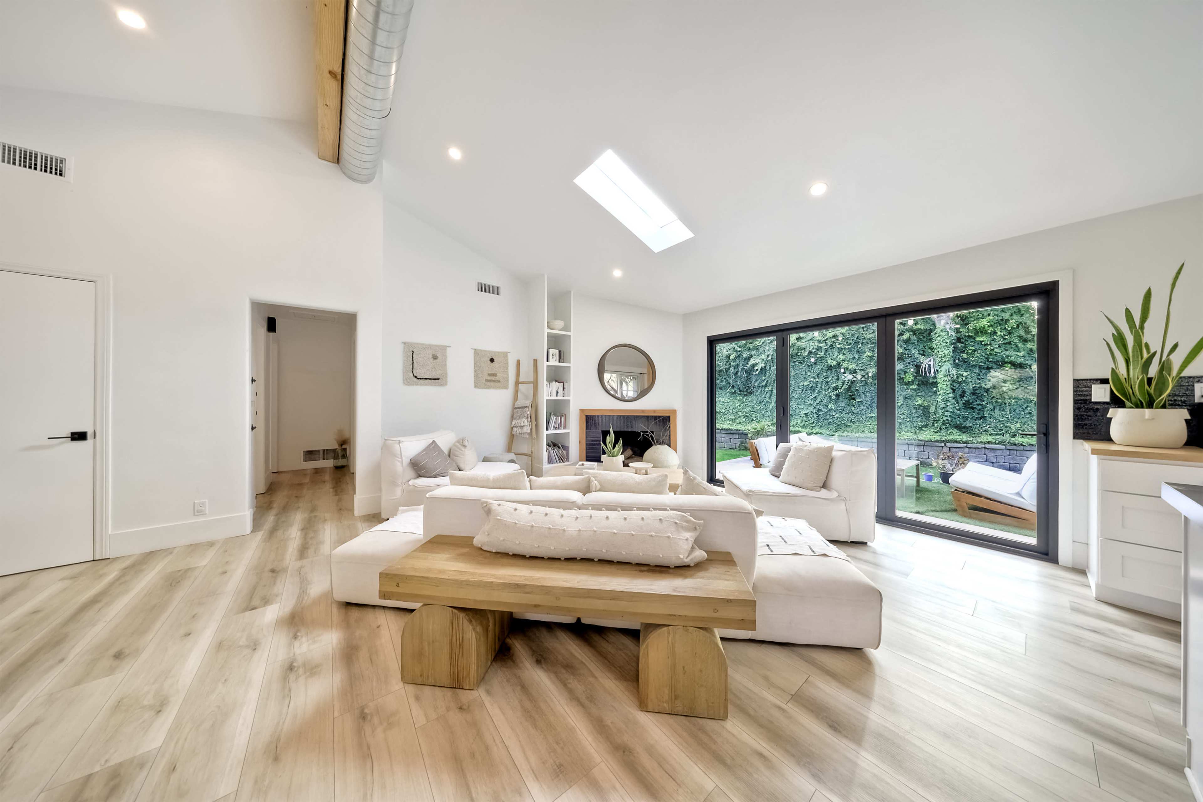 The image shows a modern living room with a light color palette, featuring a large coffee table, sectional seating, and large windows overlooking a garden.