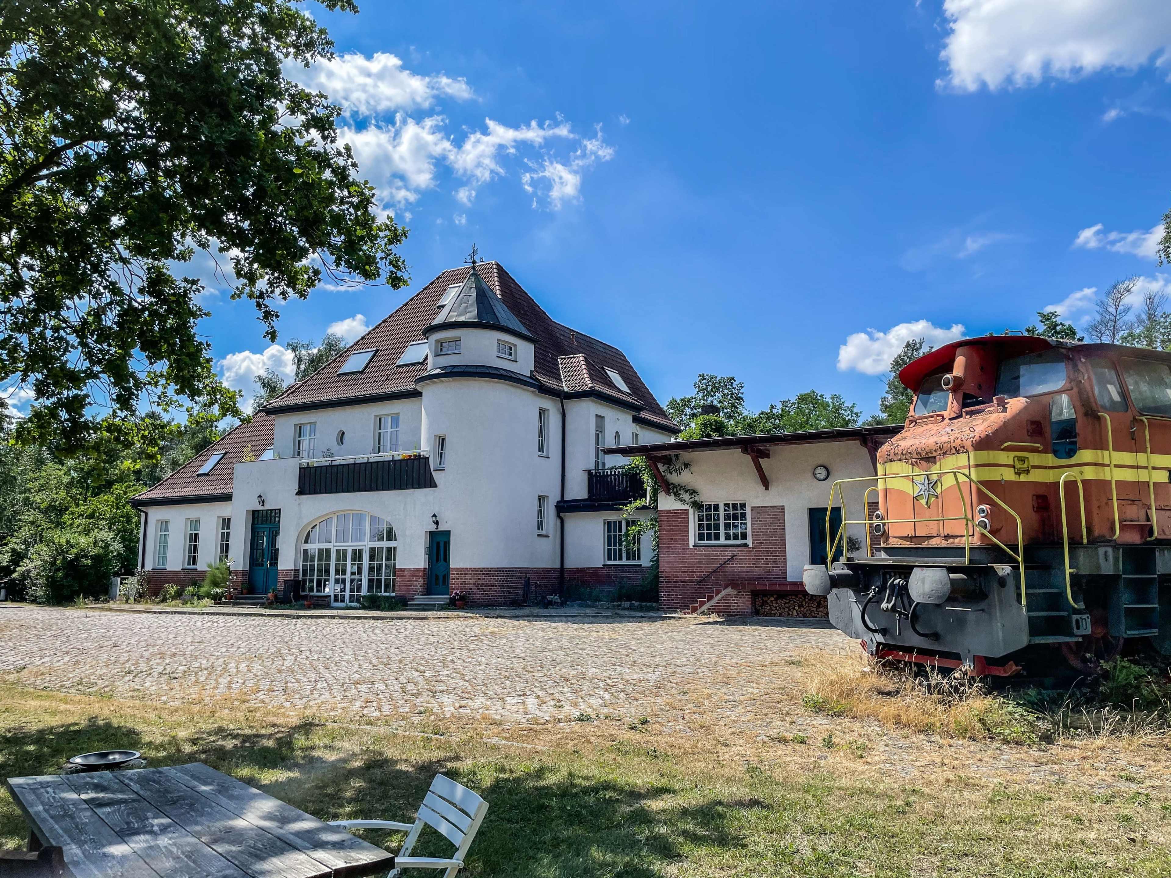 A large, white, three-story building adjacent to a parked train locomotive on a gravel area under a blue sky with scattered clouds.