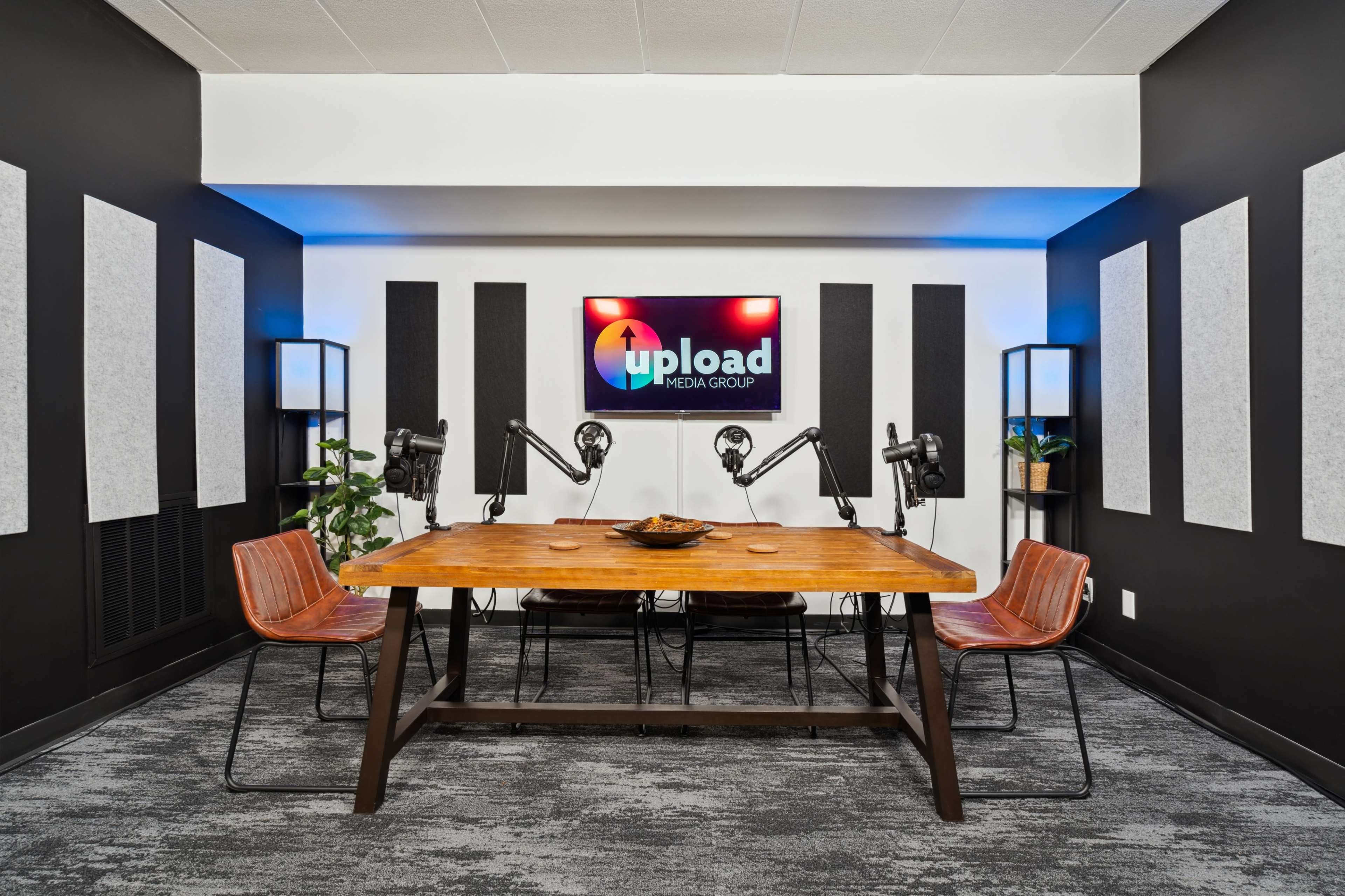 A modern conference room features a wooden table with two chairs, flanked by sound-absorbing panels and illuminated by wall-mounted lights, with a television displaying the logo of Upload Media Group.