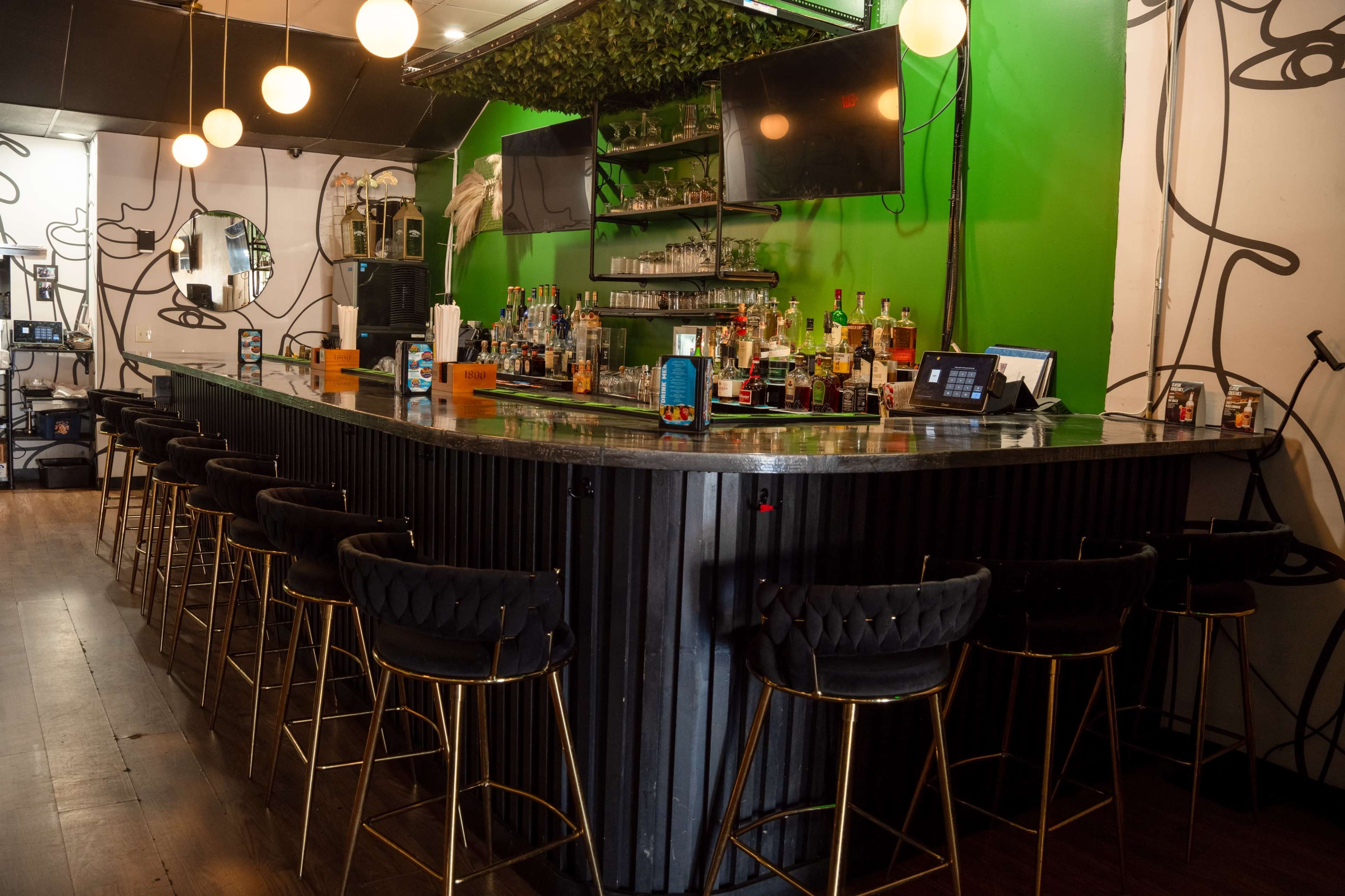 The image shows a modern bar with a dark counter, high stools, a selection of bottles on shelves, and green walls, featuring decorative elements and ambient lighting.