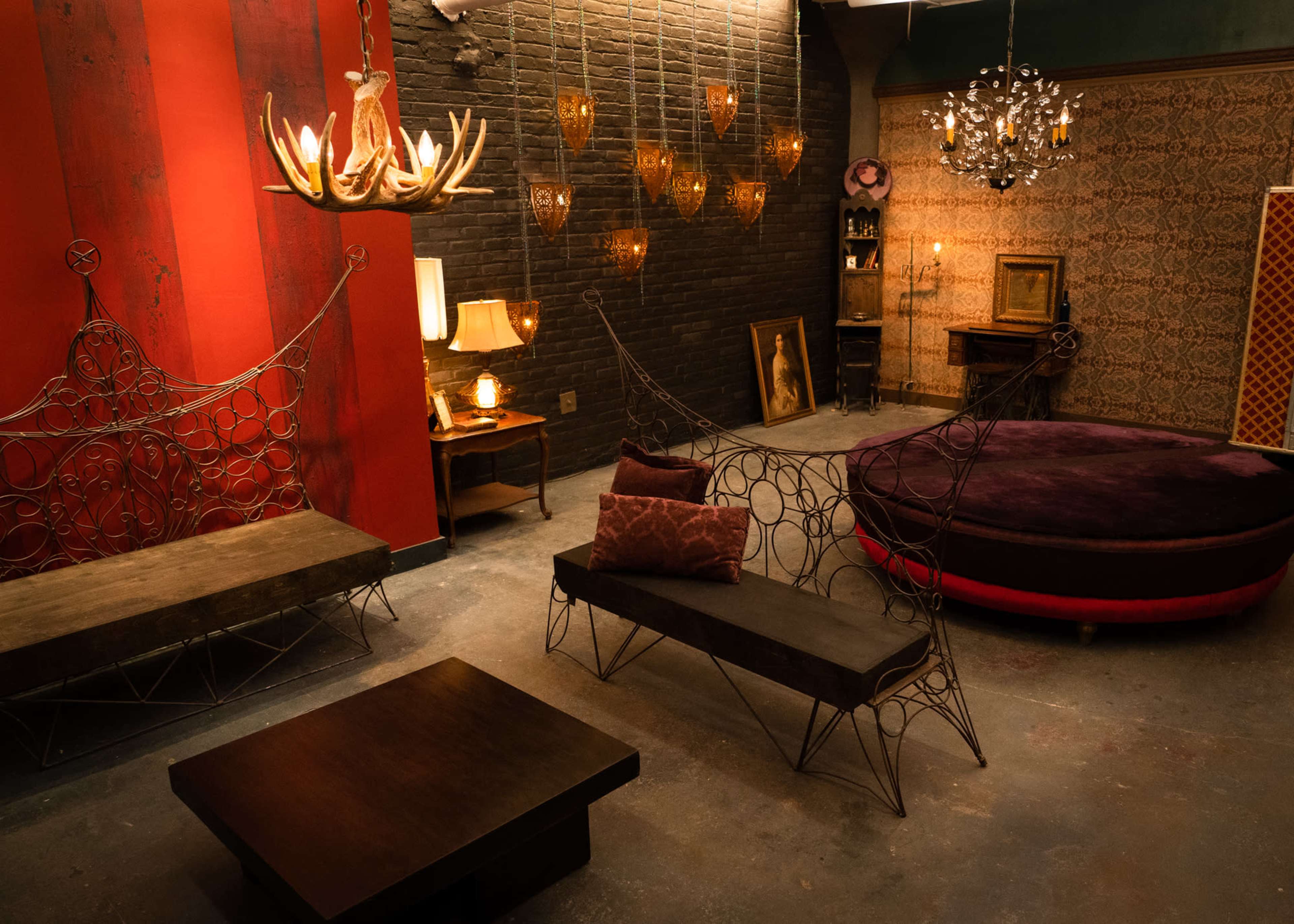 The image shows a stylish interior with various seating options, including metal benches and a round velvet couch, illuminated by distinctive light fixtures against a backdrop of red and dark walls.