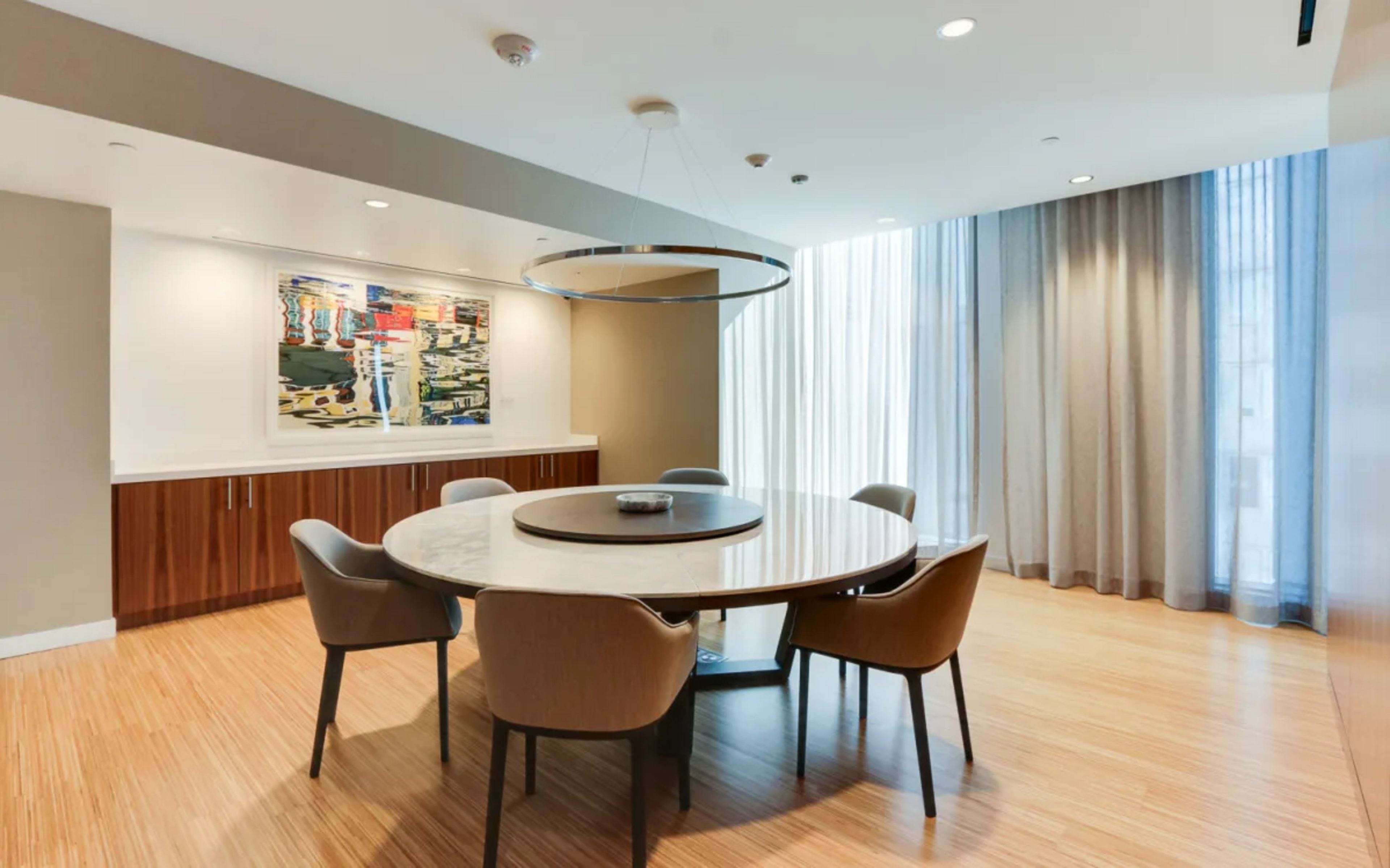 A round dining table with six chairs is centered in a light-filled room featuring a large window with sheer curtains and a modern artwork on the wall.