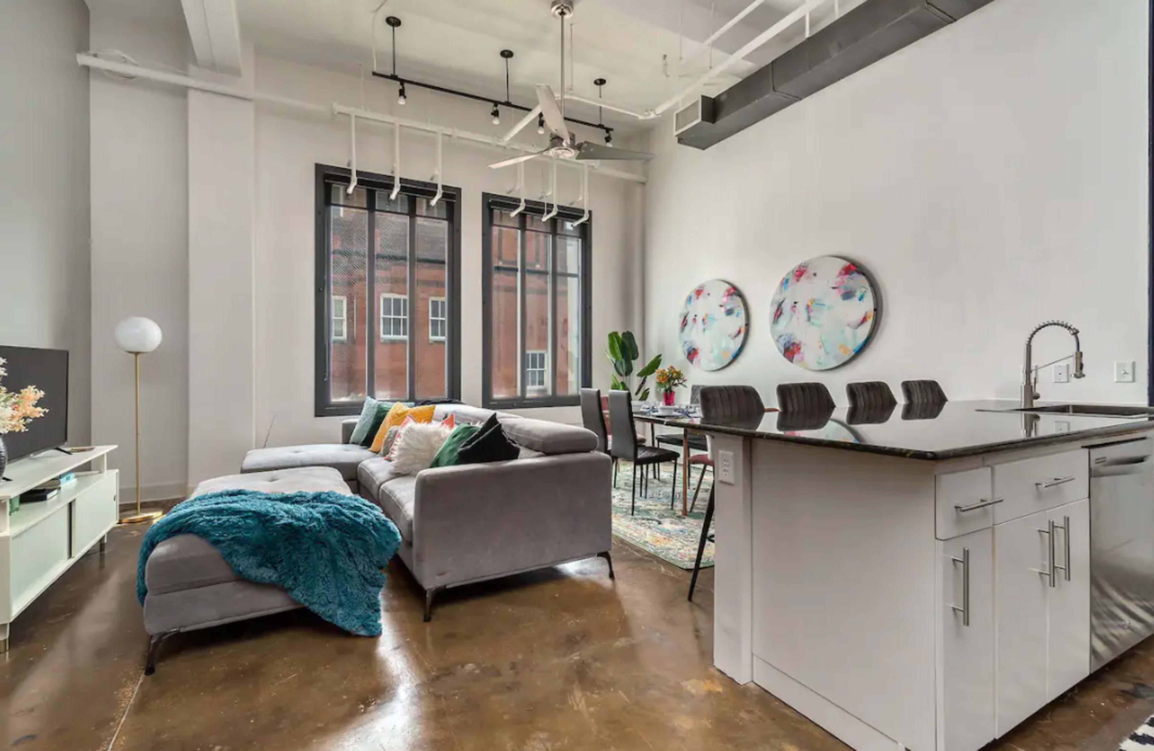 A modern living space featuring a gray sectional sofa, a dining area with a table and chairs, large windows, and abstract artwork on the walls.