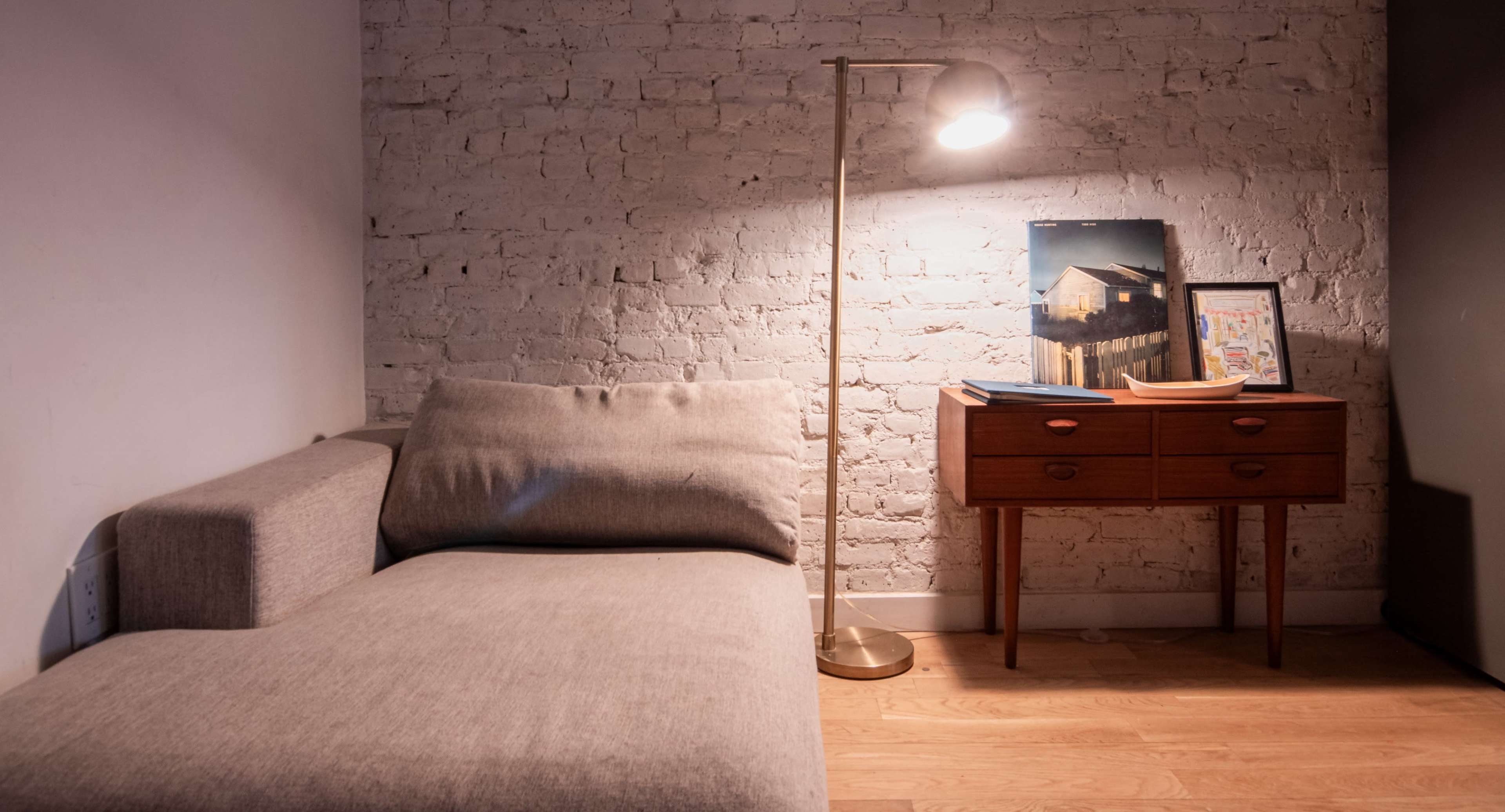 A modern living space features a gray couch, a wooden side table with decorative items, and a floor lamp illuminating the corner against a brick wall.